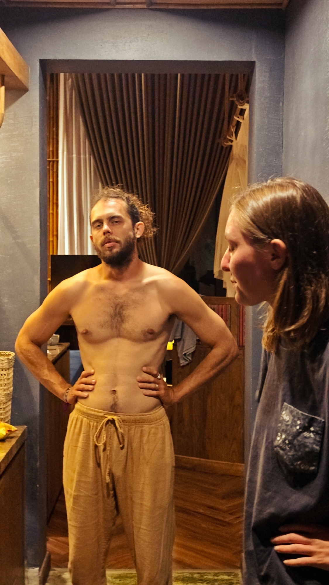 The scene is set indoors in Mu Cang Chai, Vietnam, featuring two individuals illuminated by warm, subdued lighting. A male subject, shirtless and wearing light brown drawstring pants, stands centrally on the left. He presents with a beard and mustache, and his dark, curly hair is gathered at the back of his head. Visible body hair covers his chest and abdomen. He stands with both hands placed on his hips, elbows outward, maintaining direct eye contact with the camera, his expression neutral.
To his right and slightly behind him, a female subject is partially visible in profile. She has long, light brown hair and wears a dark-colored, short-sleeved t-shirt featuring a distinct patterned patch pocket on the left chest area. Her right hand rests on her hip. She is oriented to the right of the frame, looking downwards and away from both the male subject and the camera.
The immediate background consists of dark blue, textured walls. Behind the male subject, a large rectangular opening, framed by dark wood, reveals brown, vertically pleated curtains. To the left of this opening, a vertical bamboo pole is visible. Beyond the opening, wooden plank flooring is present, appearing to be arranged diagonally or in a herringbone pattern. At the top of the frame, horizontal wooden ceiling beams or accents are visible. On the far left, a light-colored woven basket sits on a wooden counter, adjacent to a yellow object. A small section of a green rug or mat is visible on the floor at the bottom center.