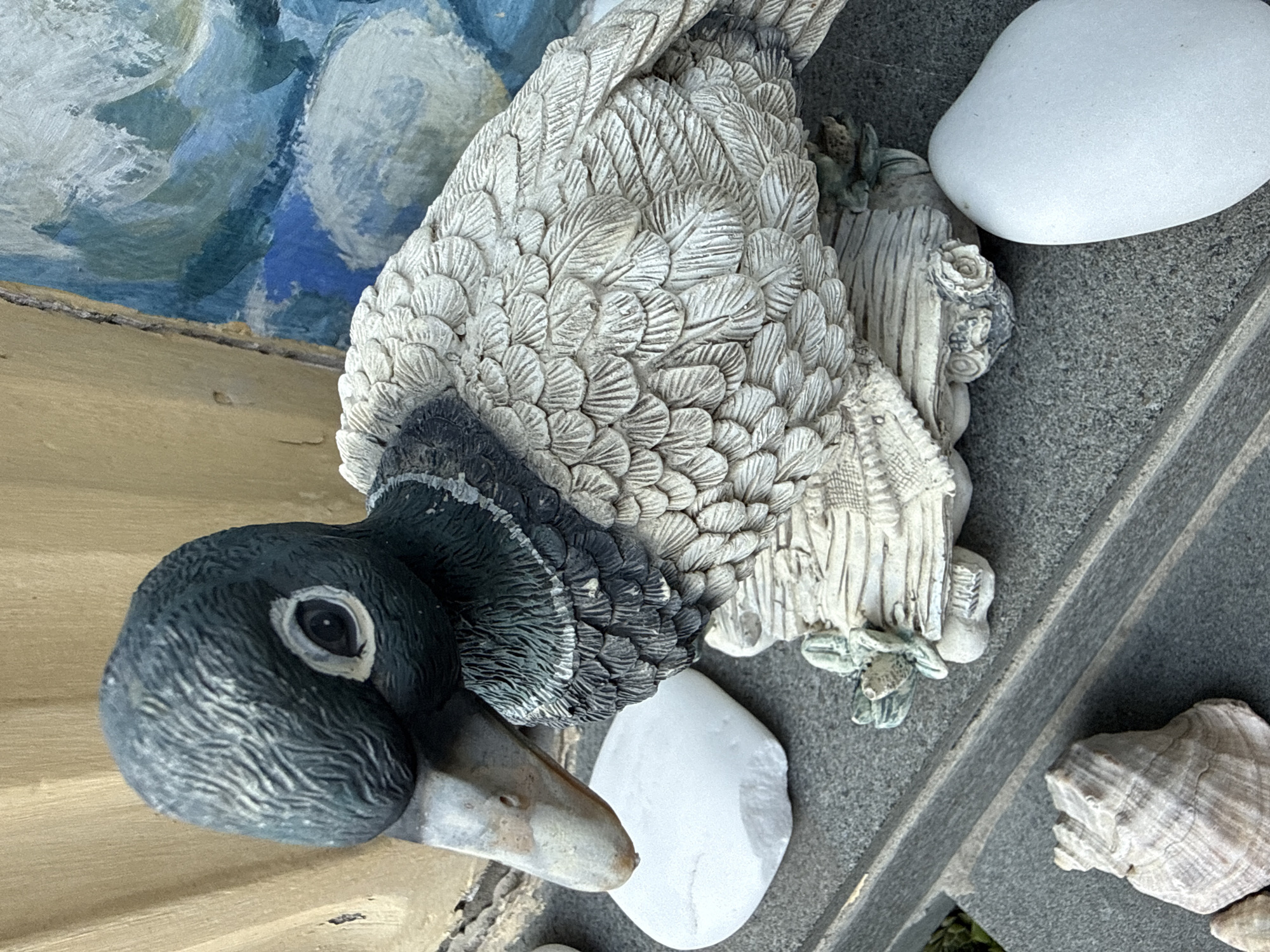 A garden figurine of a duck sits on a ledge. The duck is made of a white and gray material, with detailed feathering and a dark green head. It is positioned in front of a wall with a painted mural of blue and white abstract shapes. In the foreground, there are several white stones and a seashell. The background wall is a textured beige color. The scene appears to be outdoors, in a decorative garden or patio setting. The location is not specified to be in Okhtyrka, Ukraine.