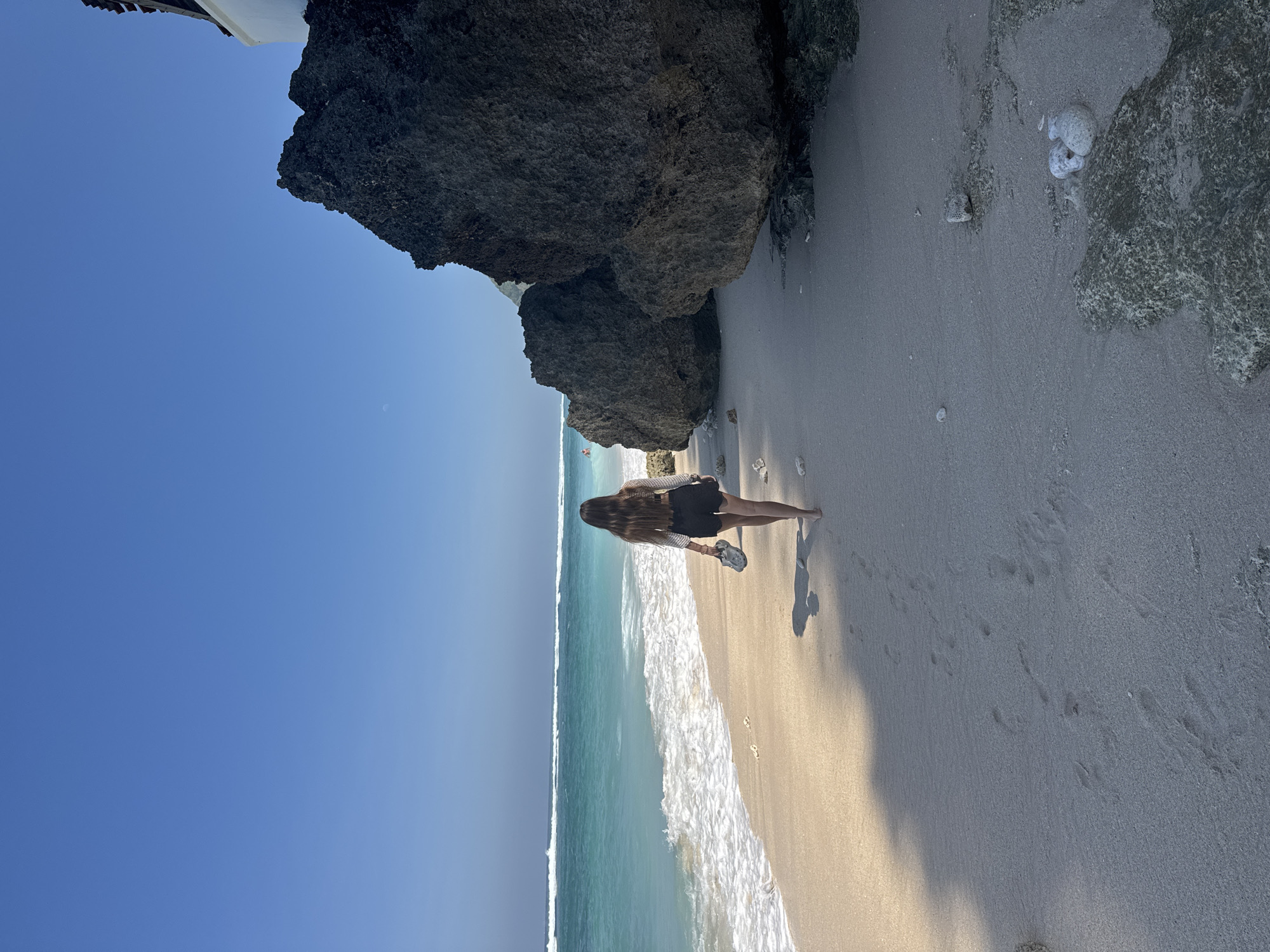 A wide-angle, sunlit outdoor shot captures a coastal scene featuring a person walking barefoot along a sandy beach towards large rock formations under a clear blue sky. The location is Unknown, Unknown.
The sky, occupying the upper portion of the frame, is a brilliant, cloudless blue, with a very faint white dot visible in the upper-left quadrant. To the left, turquoise ocean waters exhibit multiple breaking waves, with a distant, indistinct human figure possibly swimming in the water further out. The sandy beach, light beige in color, stretches from the foreground into the midground, showing a gradient from wet at the waterline to drier further up. Numerous footprints are visible in the sand, trailing away from the viewer. Small fragments of white or light-colored coral and pebbles are scattered along the beach, particularly near the waterline and the rock bases.
On the right side of the image, massive dark grey to black rock formations dominate, partially covered at their bases with lighter, textured stone or coral. A white architectural structure is partially visible behind the largest rock in the upper-right corner.
A lone individual, seen from behind, walks from the midground towards the mid-right, roughly parallel to the waterline. The person has long, straight brown hair and is wearing dark shorts and a light-colored, translucent long-sleeved top over a dark garment. They are barefoot, carrying a pair of light grey or white sandals in their left hand. A distinct shadow, consistent with the person's form, is cast on the sand to their left, indicating the sun is positioned to the right and slightly behind them.