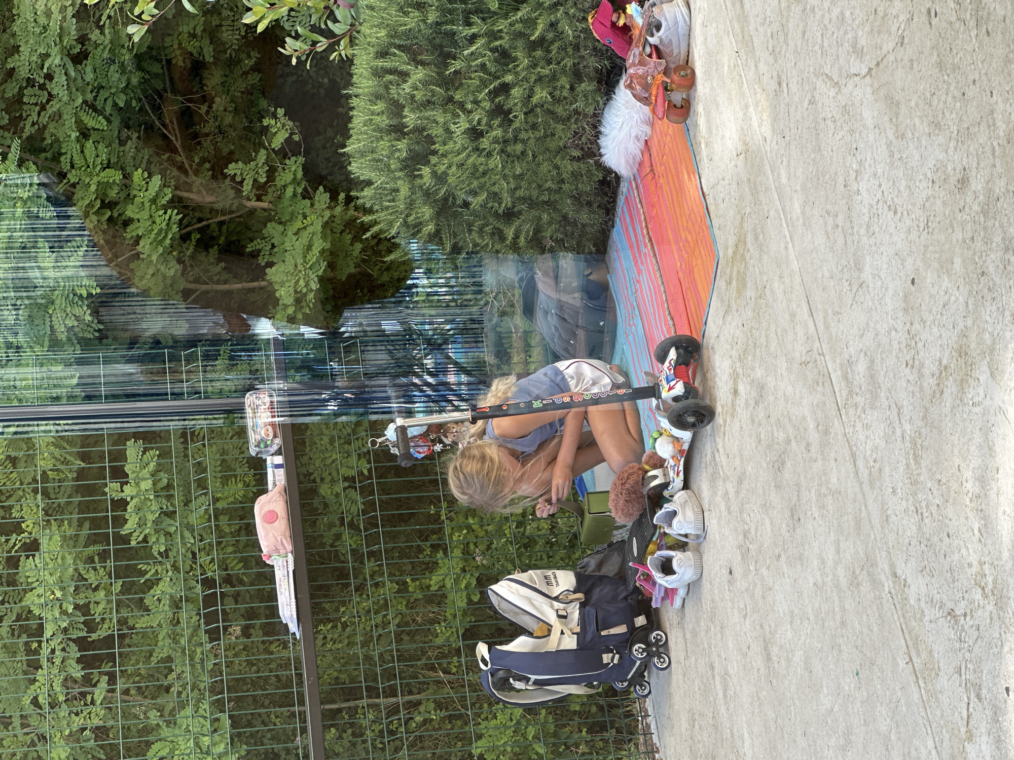 A young blonde girl with her hair pulled back is kneeling on a colorful striped mat next to a dark green bush and a metal fence. She is wearing a light blue tank top and white shorts. Her attention is focused on something in her hands. In front of her is a black scooter with a multi-colored design, a dark backpack with wheels, several pairs of shoes, a fluffy brown stuffed animal, and a skateboard. A shelf attached to the fence behind her holds a pink pouch and a clear container with pens. The scene appears to be an outdoor area, possibly a backyard or park, in Boreti, Montenegro.