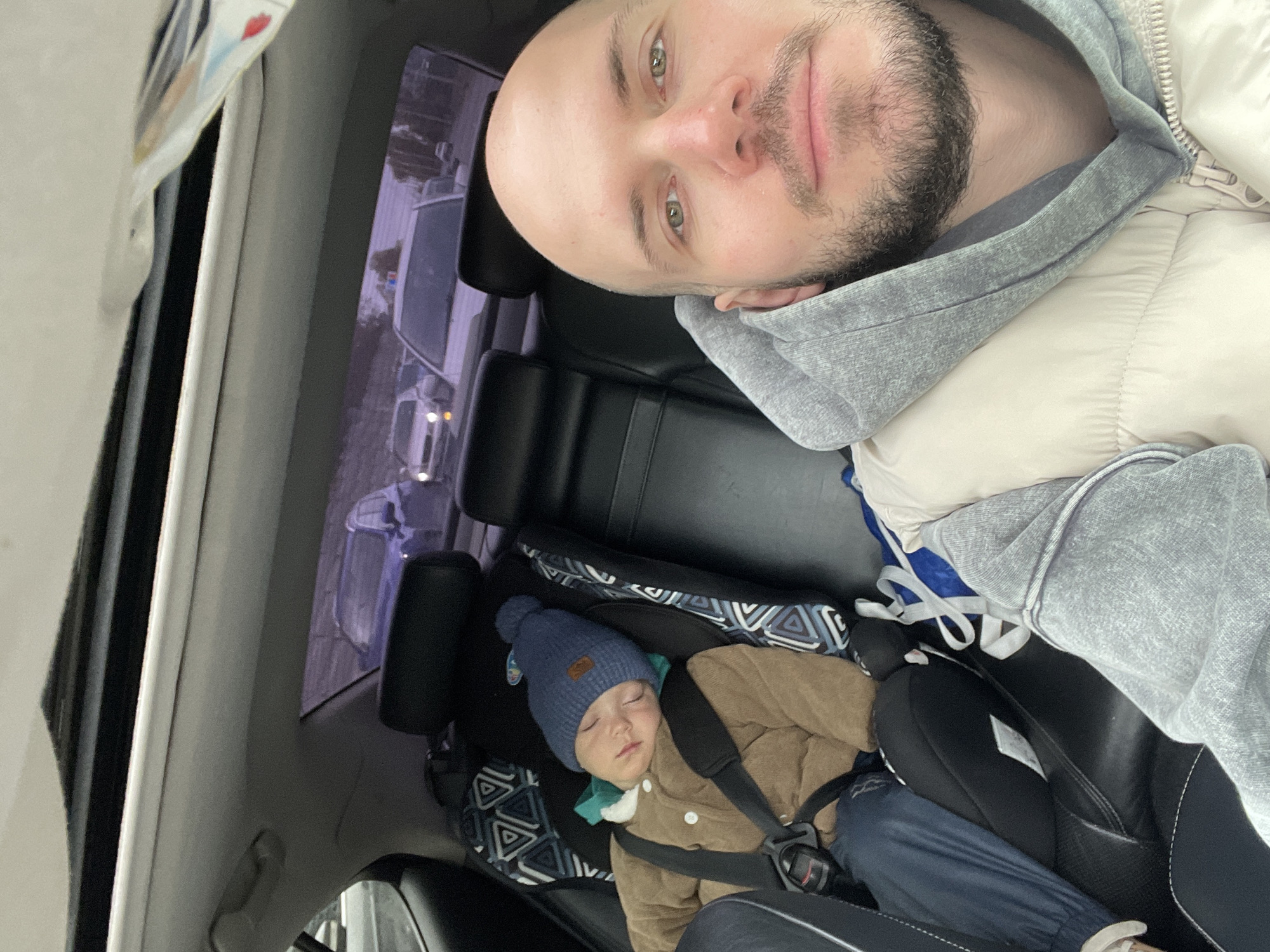 The image depicts a man and a child inside a car. The man, with a bald head and beard, is taking a selfie. He is wearing a gray hoodie and a light-colored jacket. The child, wearing a blue hat and a brown jacket, is asleep in a car seat. The car appears to have a sunroof. The reflection in the sunroof shows buildings, suggesting the car is in an urban area. Although I lack the ability to pinpoint a precise location within Krasnoyarsk, Russia, the presence of structures indicates an urban or suburban environment.
