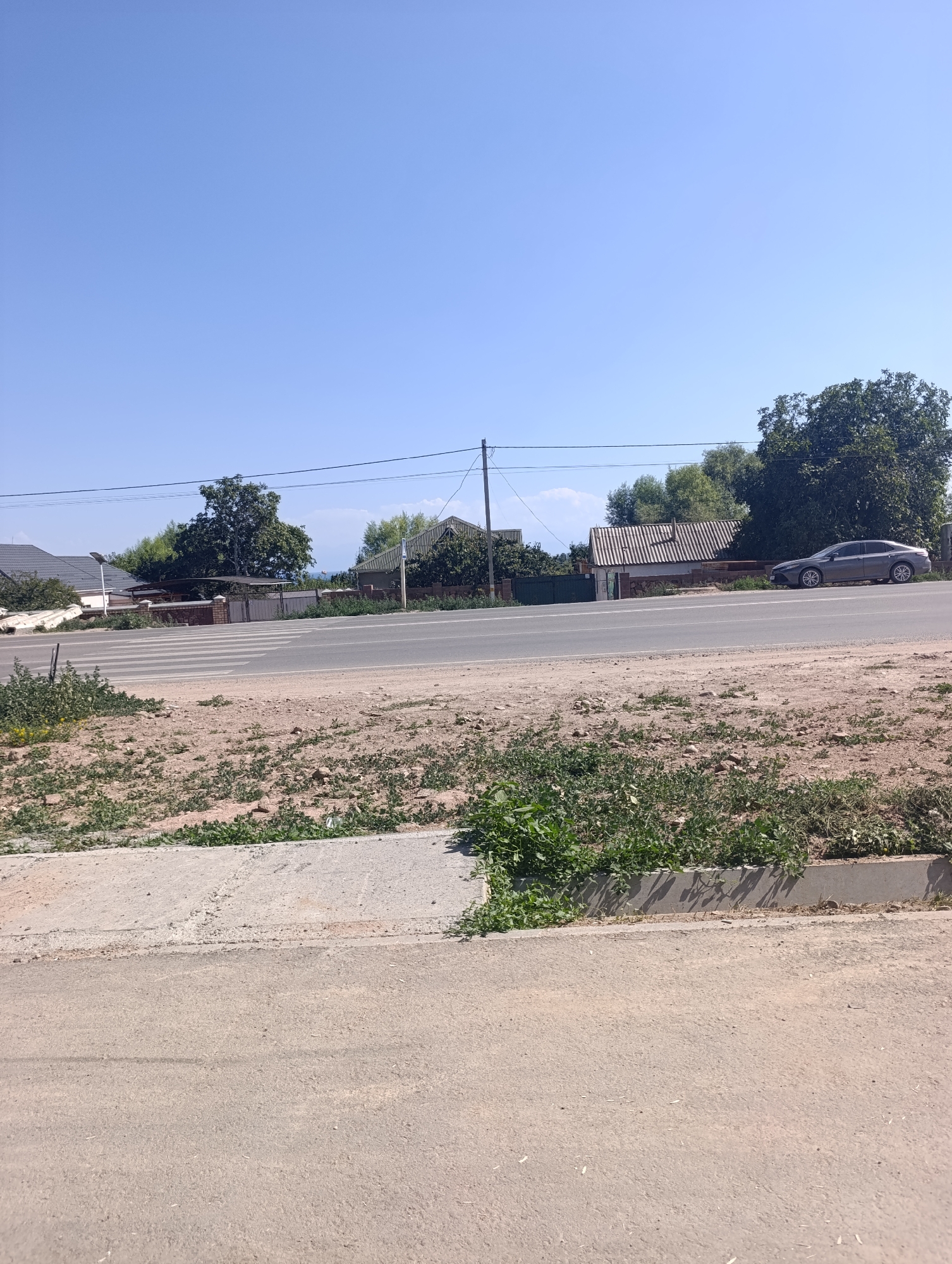 A street scene in Bosteri, Kyrgyzstan, on a clear, sunny day. The foreground shows a paved road with a concrete curb and a patch of sparse vegetation. A crosswalk is visible on the road. In the mid-ground, a line of houses and trees runs along the other side of the road. One house has a corrugated metal roof. A dark-colored sedan is parked on the side of the road in front of one of the houses. Power lines are strung across the scene. The background consists of a bright blue sky and distant hills. There are no people or animals visible in the image.