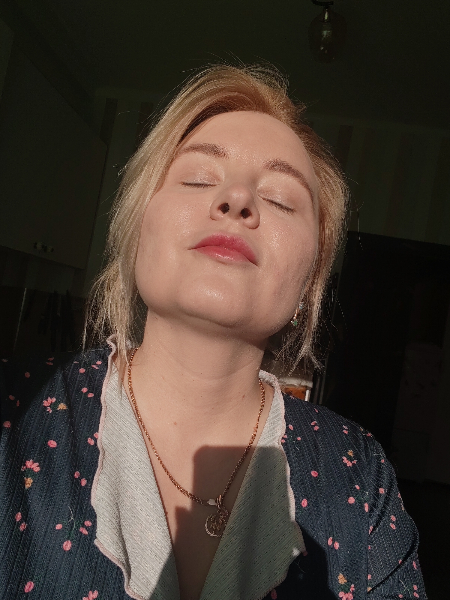 A blonde woman with fair skin is centered in a close-up shot, her eyes closed and face tilted upwards, suggesting she is basking in sunlight. She has a gold necklace and earrings. She is wearing a floral pattern blue shirt with a ruffled white collar. The background appears to be an interior space with dark walls, possibly a room in a home or apartment.
Based on the light fixture and cabinet design, the scene is likely taking place indoors, within a private residence.
There is no specific information that places the image in Izhevsk, Russia.