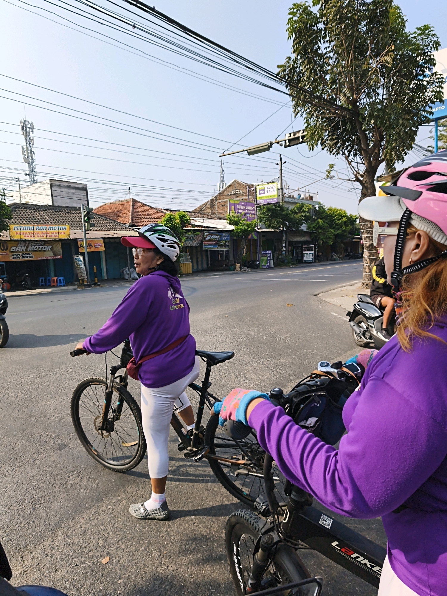 Two women are cycling on a road in the city of Surakarta, Indonesia. The woman in the foreground is wearing a pink helmet, a purple long-sleeved shirt, and white leggings. She is holding onto the handlebars of her bicycle, which has a black frame. The woman behind her is wearing a white and green helmet, a pink cap, a purple long-sleeved shirt, and white leggings. She is also on her bicycle. The road is paved and there are buildings and trees on either side. There are also power lines overhead. The time of day appears to be late afternoon, with the sun casting long shadows.