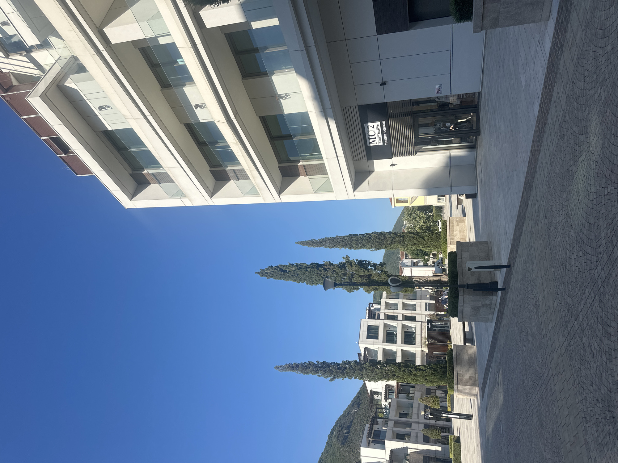 The image depicts an outdoor scene in the city of Kumbor, Montenegro, under a clear blue sky. In the foreground, a paved courtyard with a patterned brick surface leads towards several modern buildings. To the right, a white multi-story building with balconies and large windows dominates the frame. A prominent black sign with white lettering reads "MC2 SAINT BARTH TENERO FASHION" above the entrance of a store within this building. To the left, other contemporary buildings with similar architectural styles are visible, some featuring balconies and glass railings. Several tall, slender cypress trees are planted in large rectangular planters, adding vertical elements to the landscape. A lamppost is situated in the middle ground, and in the background, green hills rise against the sky. The scene appears quiet and well-maintained, suggesting a commercial or residential area. No specific actions or interactions of people are clearly discernible.