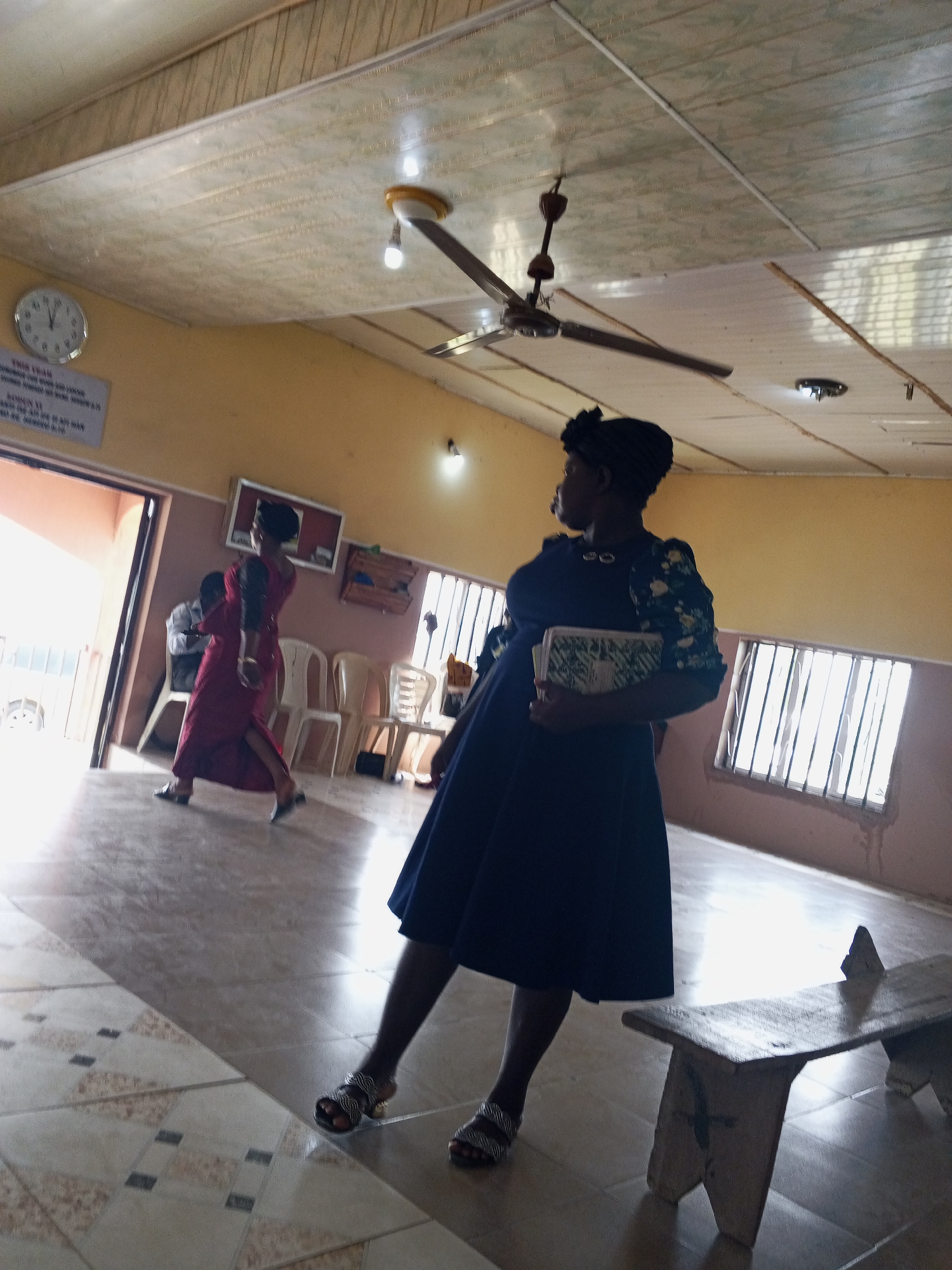 The image shows an interior space in Zango-ndaji, Nigeria, possibly a church or meeting hall. A woman in a dark blue dress and headwrap stands in the foreground, holding a book and looking to the left. Another woman in a red dress is walking through an open doorway. Chairs are lined up against a wall with windows. A wooden bench is in the right foreground. The ceiling has a fan and light fixtures. A clock and sign are visible on the wall near the door. The floor is tiled.