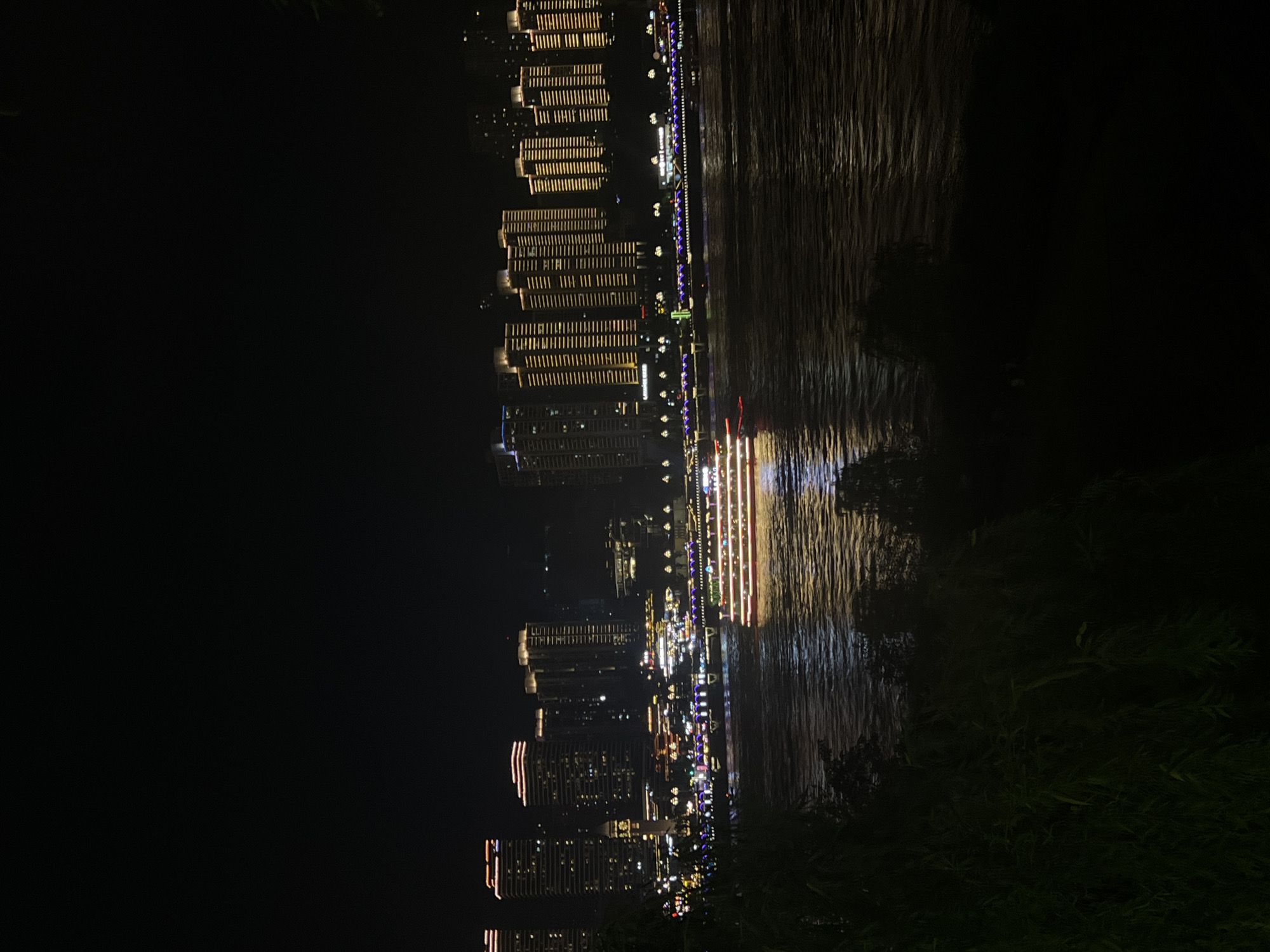 The image depicts a nighttime scene along a river in Jiangbei City Street, China. A large, illuminated cruise ship with multiple decks and red and white lights is actively moving on the dark water, casting reflections. Across the river, a city skyline of tall buildings is visible, many of which are lit up with rows of windows and decorative lights, predominantly in warm yellow and orange hues. Some of the buildings have distinctive architectural features, such as stepped roofs or illuminated bands. Along the riverfront, there are more lights from what appear to be buildings, possibly a promenade or other structures. The water surface is rippled, reflecting the artificial lights from the city and the ship. In the foreground, dark silhouettes of trees and foliage obscure the lower portion of the scene, creating a framing effect. The overall atmosphere is one of urban nighttime activity and illumination.