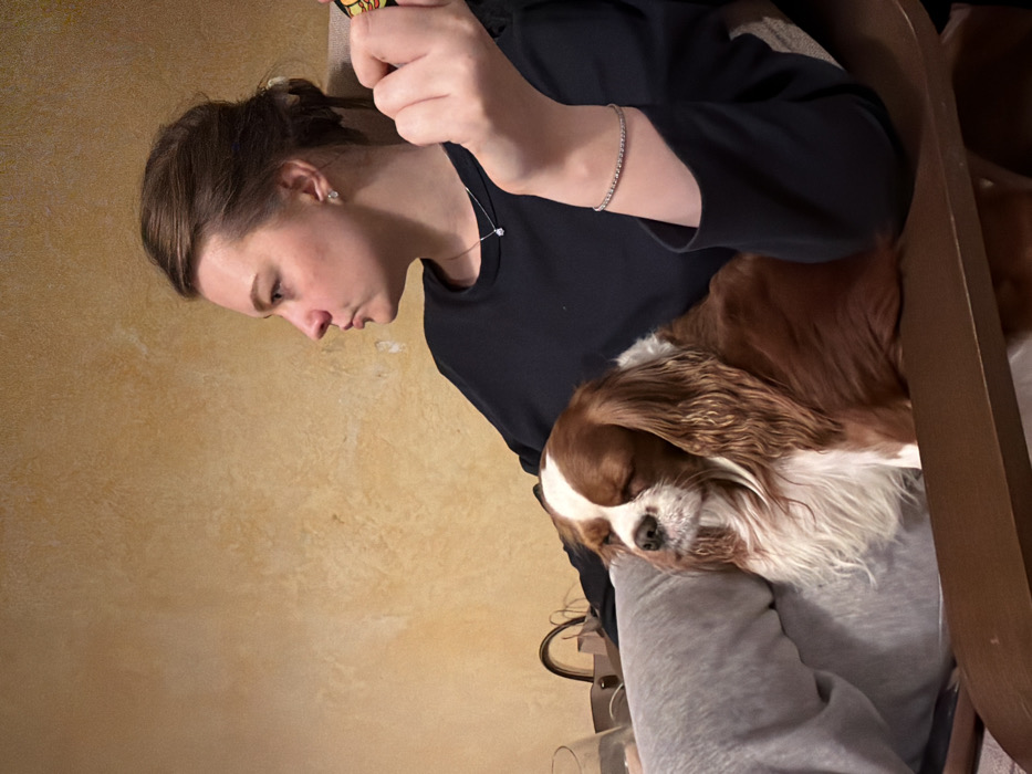 A woman sits with a Cavalier King Charles Spaniel nestled on her lap, appearing to be asleep. She is wearing a black long-sleeved shirt and a silver bracelet. Her hair is pulled back in a bun, and she has on a necklace and earrings. A wooden chair can be seen to the right. The wall in the background is a textured light brown color.