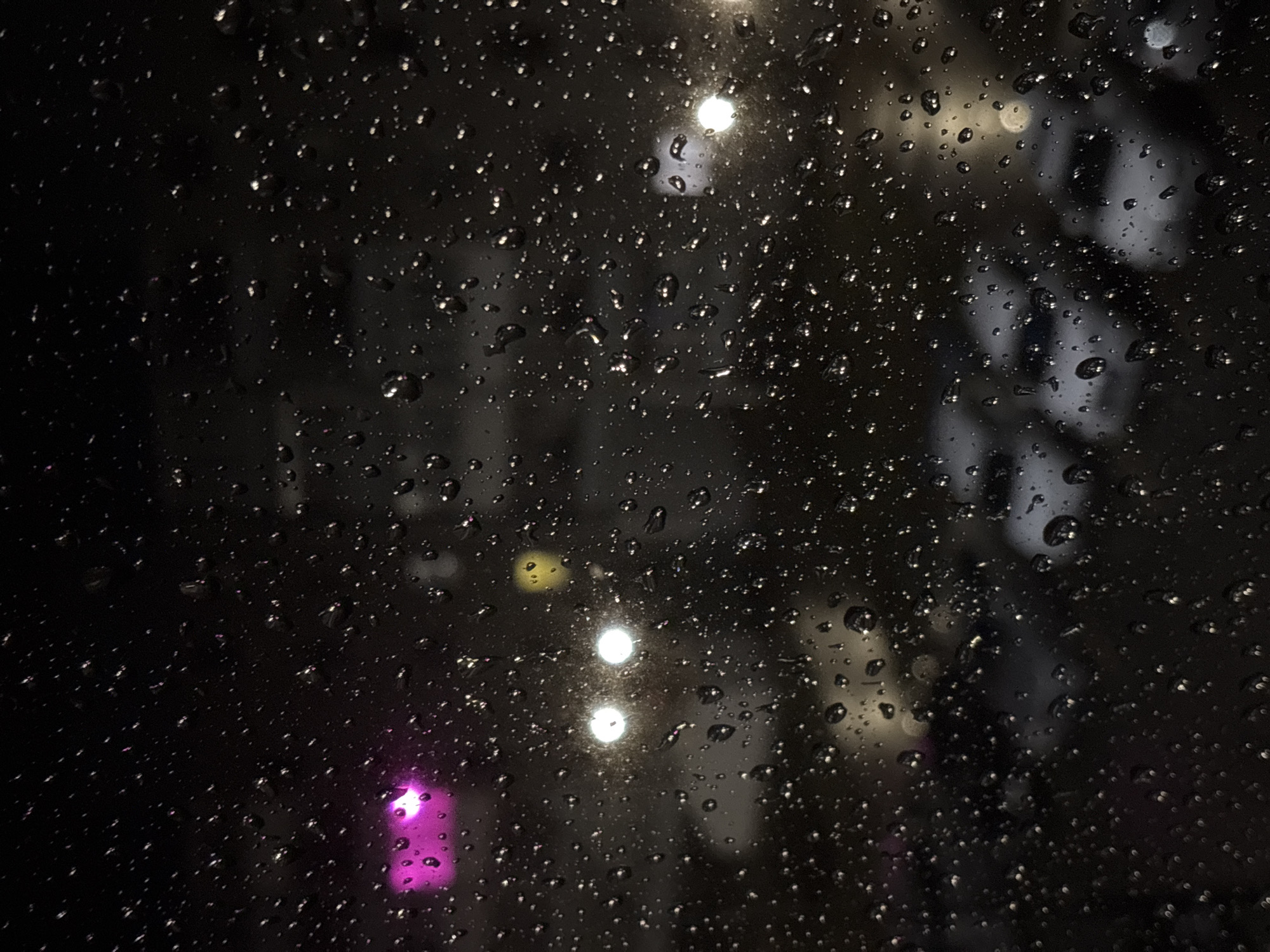 The image displays a close-up, abstract view of a transparent surface covered in numerous water droplets, characteristic of a window or glass pane during or after rain. The droplets vary in size and create a textured, shimmering effect across the dark foreground, with many reflecting light to appear as bright, glistening points.
The background is dark and heavily out-of-focus, strongly indicating a nighttime scene. Through the blurred backdrop, several distinct light sources are visible. There are multiple bright white circular lights, appearing as diffused orbs, scattered across the mid-ground. A prominent magenta or purple light glows in the lower-left portion of the image, while a subtle yellow light blur can be seen towards the upper-center. Beyond these lights, indistinct, shadowy shapes suggest distant objects or structures in an urban or semi-urban environment.
No specific activity or event is depicted, rather it is a static, atmospheric shot focusing on the interplay of light and water on glass. The weather is clearly rainy or recently rained. There are no people, recognizable objects, or visible text in the image. The scene evokes a moody, contemplative atmosphere often associated with rainy nights. The implied location is Марковский, Russia.