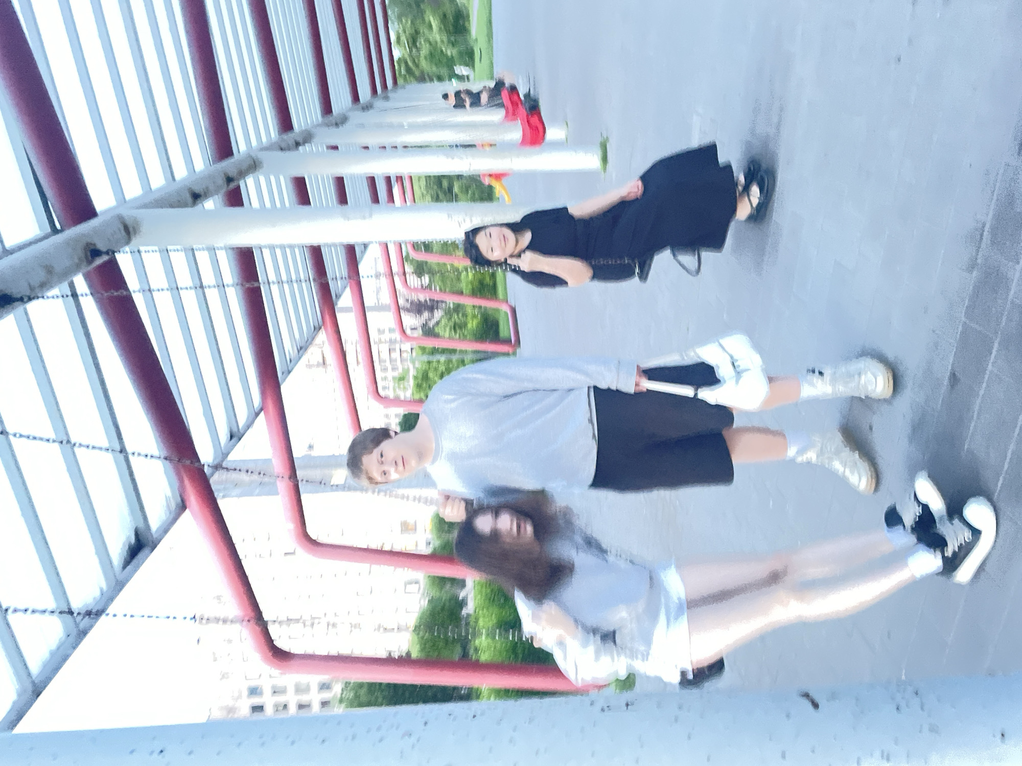 In Sinyavino, Russia, three individuals are present at an outdoor playground with a covered seating area. In the foreground left, a female with long dark hair is leaning forward and appears to be speaking or reacting. She is wearing a light gray sweatshirt, a white skirt, sheer white stockings, and black platform sneakers. To her right, a male stands looking towards the camera, wearing a light gray long-sleeved shirt and dark shorts. He is holding onto a chain from the playground structure. In the mid-ground right, a female is seated on a swing, wearing a black dress and black platform shoes. She is looking at the camera and smiling. The playground features red metal beams and white pillars supporting a corrugated roof. Buildings and trees are visible in the background. The ground is paved.
