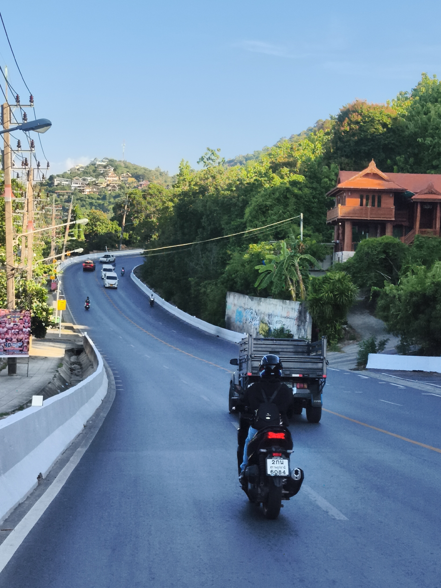 This is a photograph taken on a road in the Ko Samui District of Thailand. The road curves through a lush, green, hilly landscape. In the foreground, a person is riding a black scooter with a license plate that reads "2คน 6084". The rider is wearing a black jacket and a helmet. Behind the scooter, a flatbed truck is also traveling on the road. The road has a yellow center line. On the left side of the road, there are utility poles and a white concrete barrier. In the background, there is a large, multi-story wooden building with a distinctive orange roof, nestled amongst the trees on a hillside. Further up the hill, several houses are visible. The sky is clear and blue. The overall impression is a sunny day on a scenic road in a tropical environment.