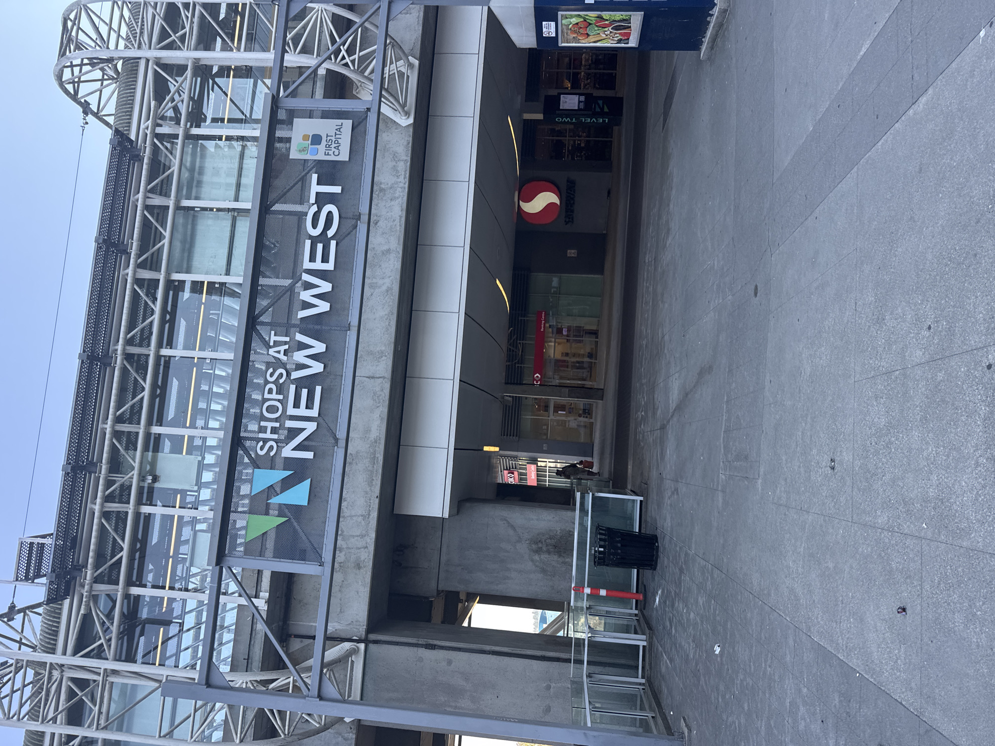 The image shows the exterior of a shopping center in New Westminster, Canada. A prominent sign above the entrance reads "SHOPS AT NEW WEST". Below the sign, there is a logo with the text "FIRST CAPITAL". The entrance to the shopping center features a Safeway store with its red and white circular logo. There are glass doors and windows leading into the stores. In the foreground, the ground is paved with concrete tiles, and there is a black trash can and a red and white traffic cone. A person is visible walking into the shopping center. The architecture of the building is modern, with a lot of concrete and metal structures, and large glass panels. The sky above is clear and blue.