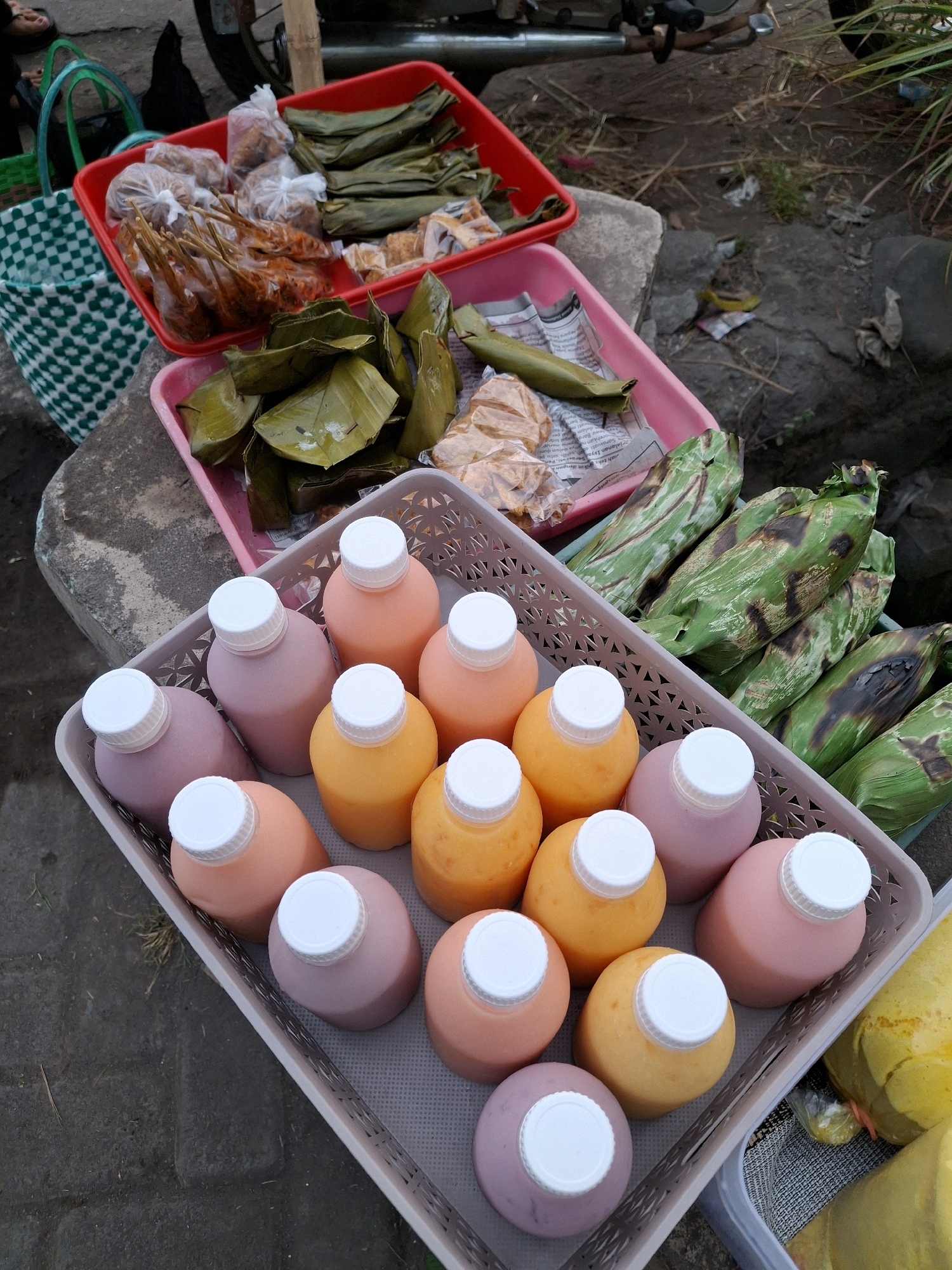 The image captures a street-side food stall in Lumajang, Indonesia, likely on a sidewalk or roadside area characterized by rough concrete and natural ground. In the foreground, a light-colored plastic basket is filled with numerous plastic bottles containing colorful beverages, some appearing to be orange and pink or purple. These bottles have white caps. To the right of this basket, another basket contains grilled food items wrapped in green leaves, with some charring visible on the leaves, suggesting they have been cooked over heat. Behind the bottles, in a red plastic tray, are various food items, some packaged in clear plastic bags and others wrapped in leaves. There are also what appear to be skewers of cooked food. A green and white checkered reusable bag is visible to the left. The overall impression is of a typical Indonesian street food vendor offering a variety of prepared foods and drinks.