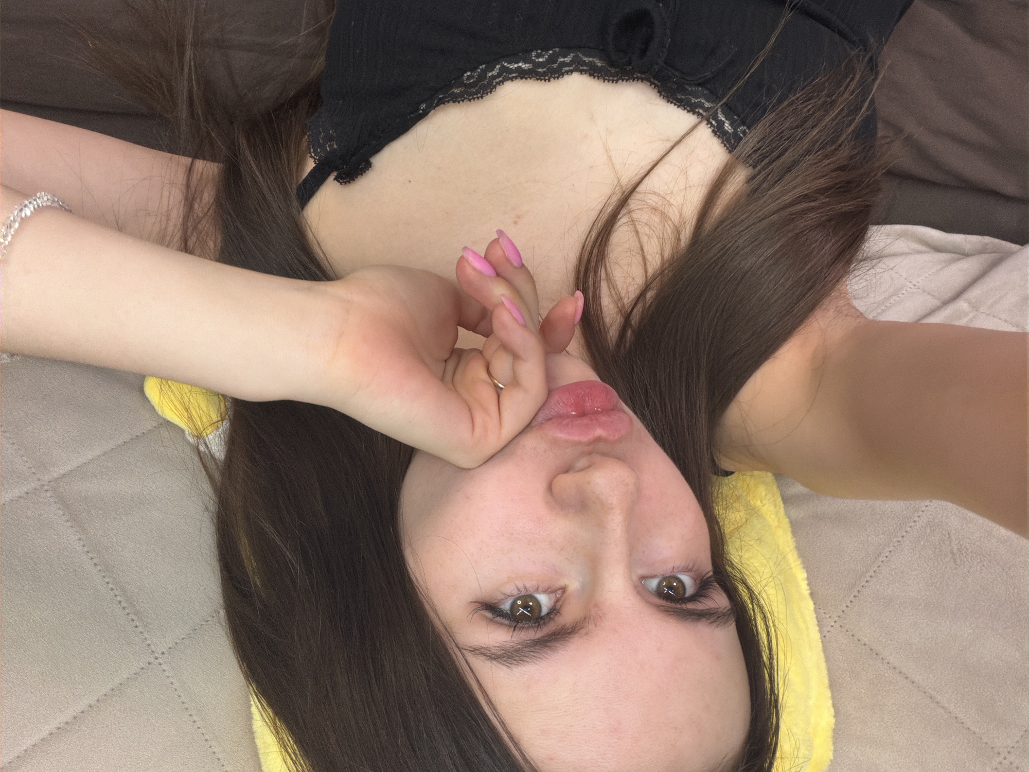 A close-up, portrait-oriented image shows a young female lying down in what appears to be an indoor residential setting. She has long, straight, dark brown hair that spreads around her head. Her complexion is fair, with visible freckles across her nose and cheeks. She has brown eyes with prominent eyelashes, looking directly at the camera. Her lips are pursed in a pout, displaying a pinkish color. Her left hand is raised, with fingers curled and pink nail polish, and a gold-toned ring is visible on one finger, resting near her chin. A silver-colored, possibly beaded, bracelet is worn on her right wrist, which is partially visible in the upper right. She is wearing a black top with lace trim along the neckline. The background features a light brown or beige quilted surface, possibly a bedspread or sofa, with a stitched diamond pattern. A bright yellow soft object, possibly a pillow, is visible beneath her head on the left side of the frame. Darker brown fabric is present on the right side, and a lighter, crumpled fabric is in the lower right corner.