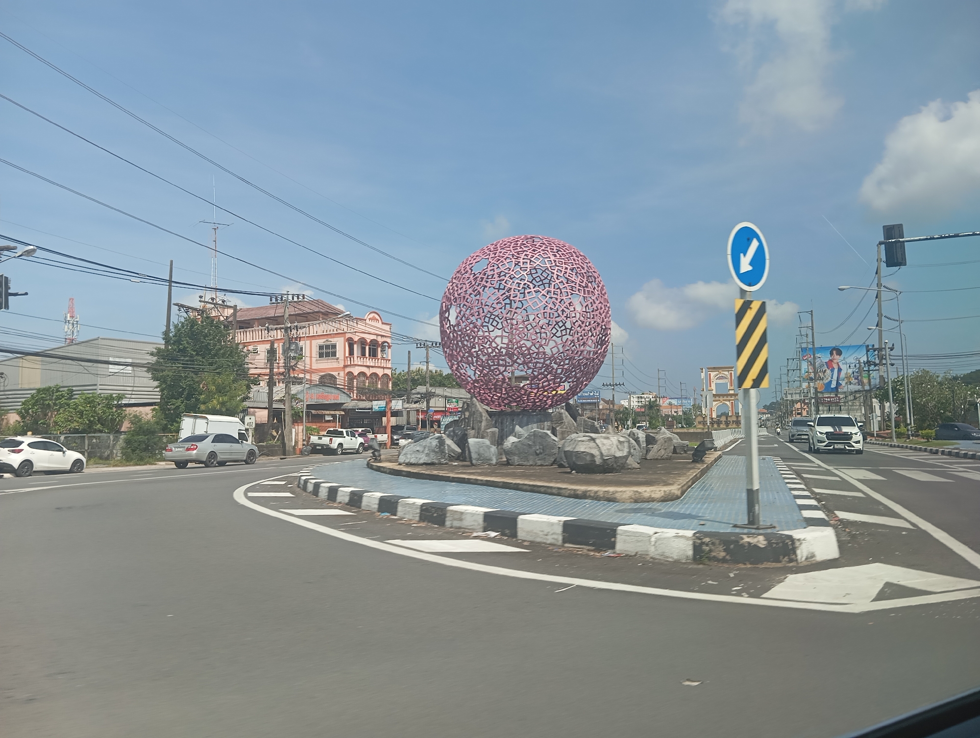 This image captures a traffic circle in Ko Kaeo, Thailand, featuring a prominent spherical sculpture constructed from pink, interconnected geometric shapes. The sculpture is surrounded by large, natural rocks and situated on a paved island with blue and white patterned tiles. The traffic circle is bordered by a curb with black and white stripes. Several vehicles are visible, including a white Mazda on the left, a silver sedan, and a white pickup truck. Traffic signs, including a blue sign with a white arrow indicating a directional change, are also present. In the background, buildings with arched windows and balconies, power lines, and streetlights line the street. A tall antenna mast is visible in the distance. The sky is a clear blue with scattered white clouds.