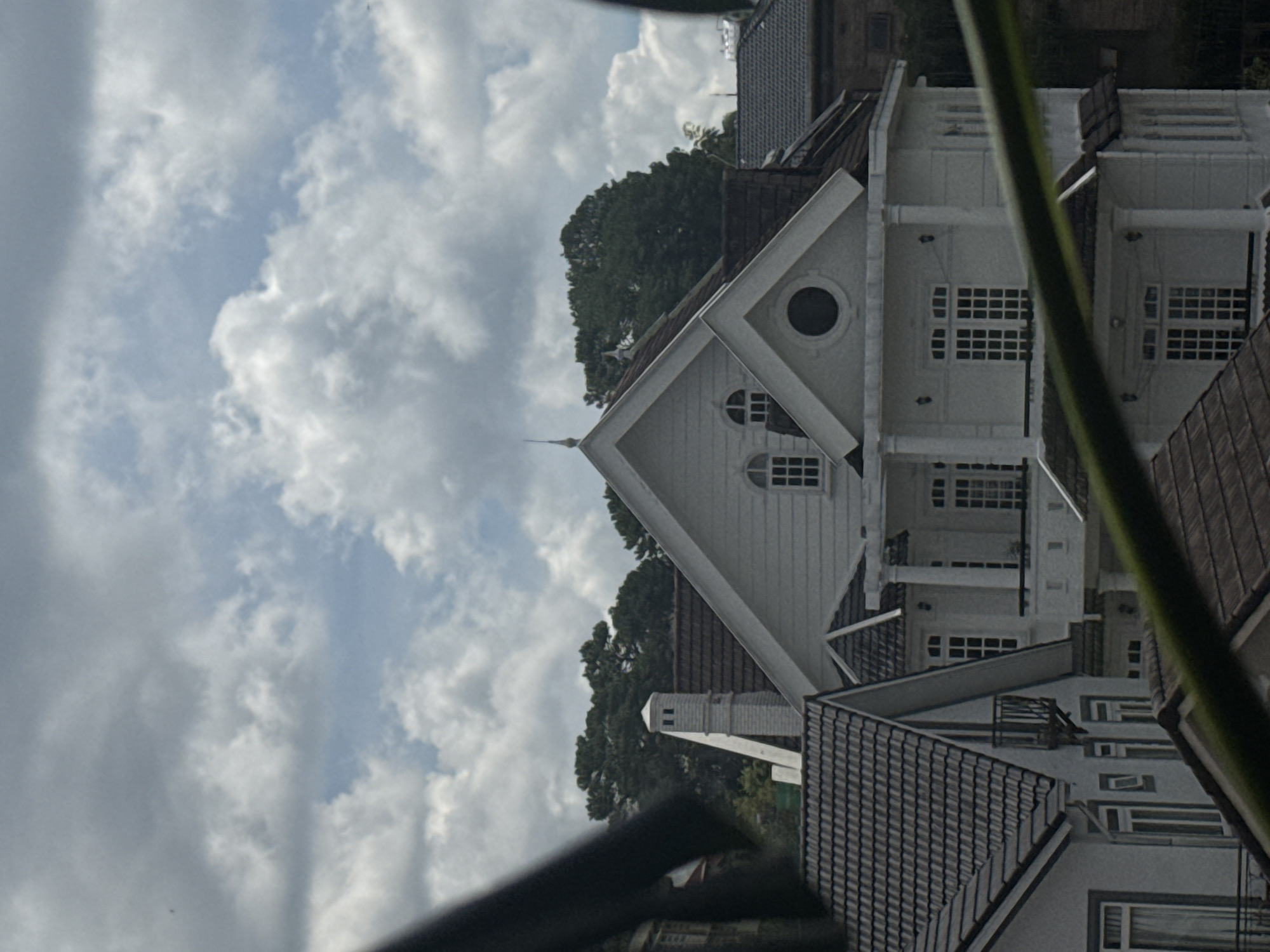 The image displays a section of Da Lat, Vietnam, characterized by multi-story houses with prominent architectural features. The sky above is filled with dramatic, voluminous clouds against a pale blue backdrop, suggesting a dynamic weather pattern. In the foreground, partially obscuring the view, are dark, out-of-focus elements that appear to be foliage or possibly a frame.
The primary focus is on a series of white, European-style houses with steep, dark-tiled roofs. One prominent house in the center features a tall, white chimney with square openings and a pointed cap, indicative of a classic design. The house has multiple windows, many with white frames and multi-pane panes, and a balcony supported by columns. To the left, another house exhibits a similar roofline and windows. To the right, a darker, brick-colored building is visible, also with a tiled roof. The surrounding area is densely populated with evergreen trees, their dark green foliage creating a stark contrast with the lighter-colored buildings and the bright sky. The overall impression is one of a residential area with distinct architectural influences, set against a backdrop of natural, if cloudy, weather.