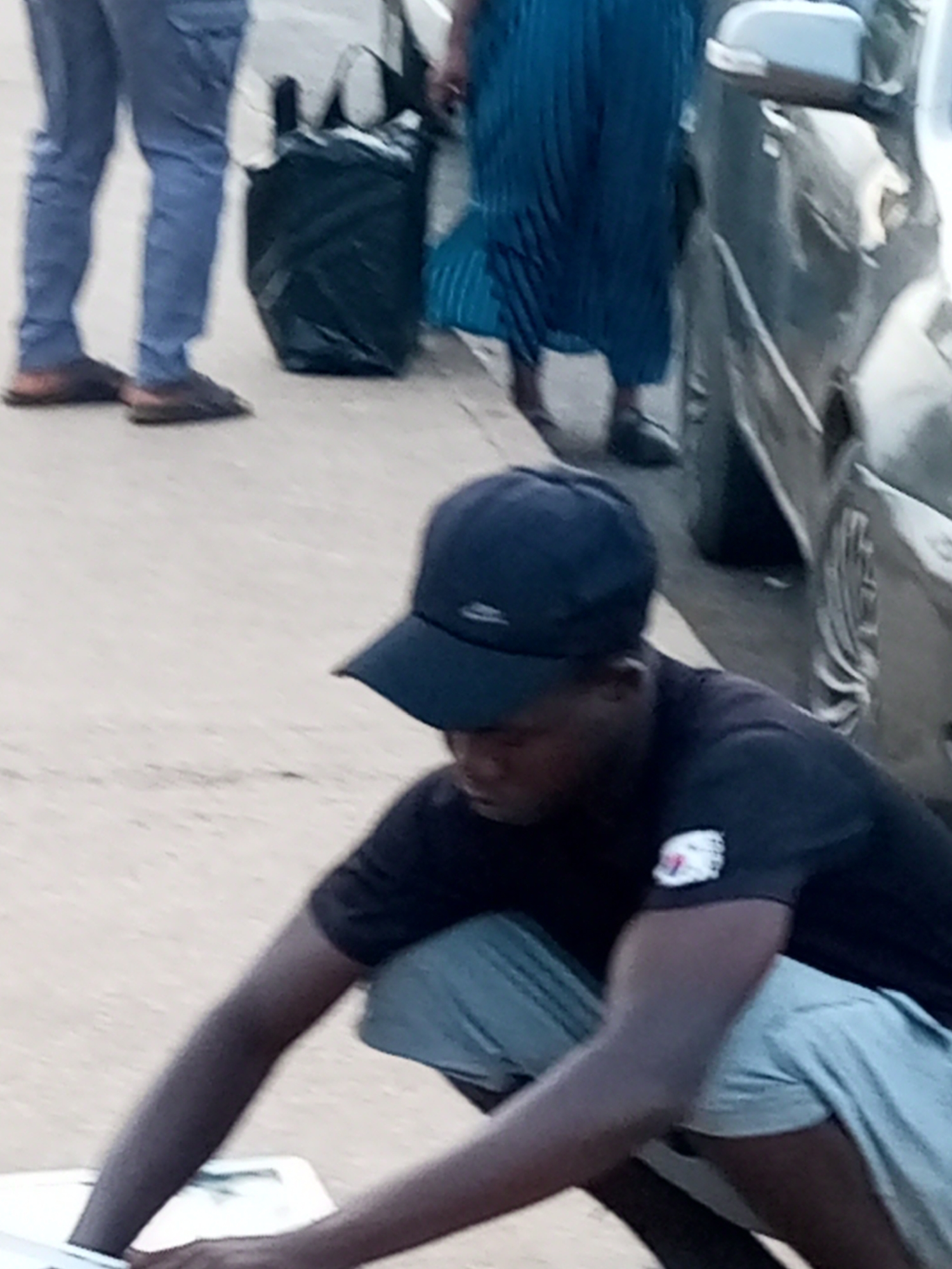 In Gombe, Nigeria, an individual wearing a dark t-shirt and a dark baseball cap is crouching down. The person's t-shirt has a patch on the left sleeve, and they are wearing light-colored shorts. In the background, another person is visible wearing blue trousers and brown shoes, standing next to a large black trash bag. A portion of a car is also visible in the background. The scene appears to be taking place outdoors on a paved surface.