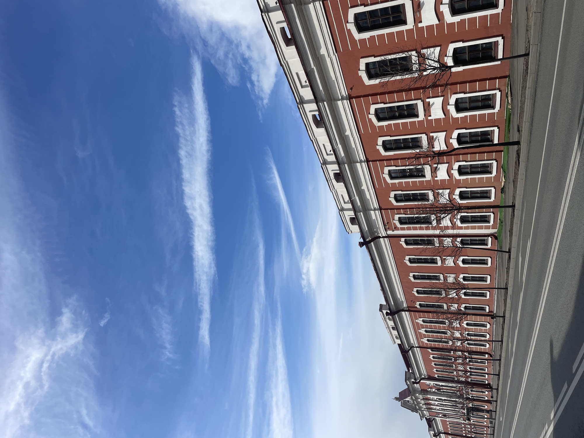 The image depicts a long, low building in Kazan, Russia, taken from an angled, street-level perspective. The building has a distinctive red facade punctuated by rows of windows with white trim, a large cornice and is bordered by a sidewalk and a two-lane road with a double white line in the middle. The sky takes up the left half of the frame, showcasing a clear blue sky with wispy, white cirrus clouds. Thin, leafless trees are planted along the sidewalk, equidistant from each other. The overall impression is one of an urban landscape on a clear, sunny day.