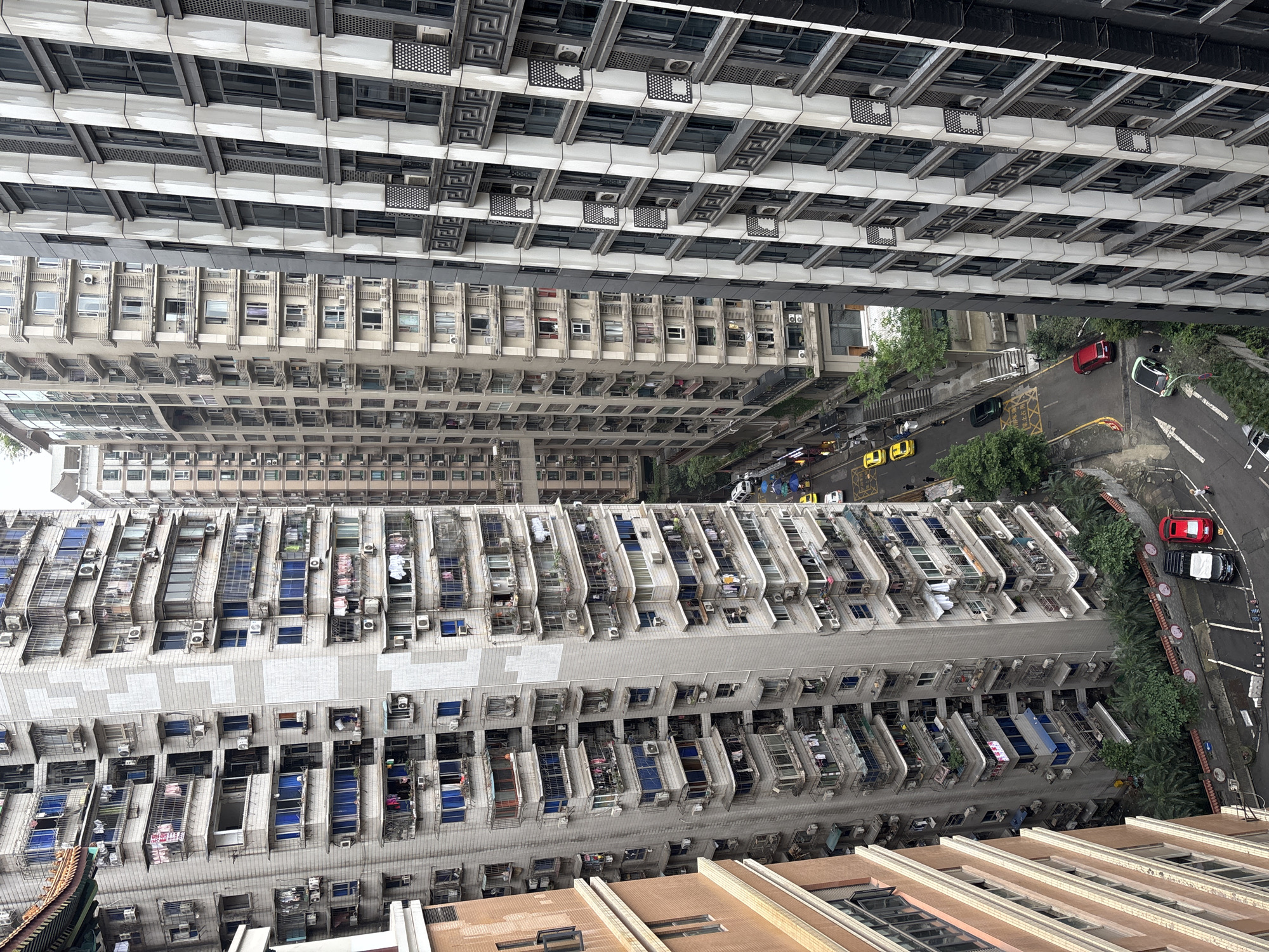 The image depicts a dense urban landscape in Yuzhong District, China, viewed from a high vantage point. Numerous tall residential buildings dominate the scene, their facades characterized by repetitive rows of windows and balconies, many equipped with air conditioning units and clothes drying racks. A street runs between some of the buildings, where a few yellow taxis and other cars are visible. The overall atmosphere suggests a densely populated residential area within a large city.