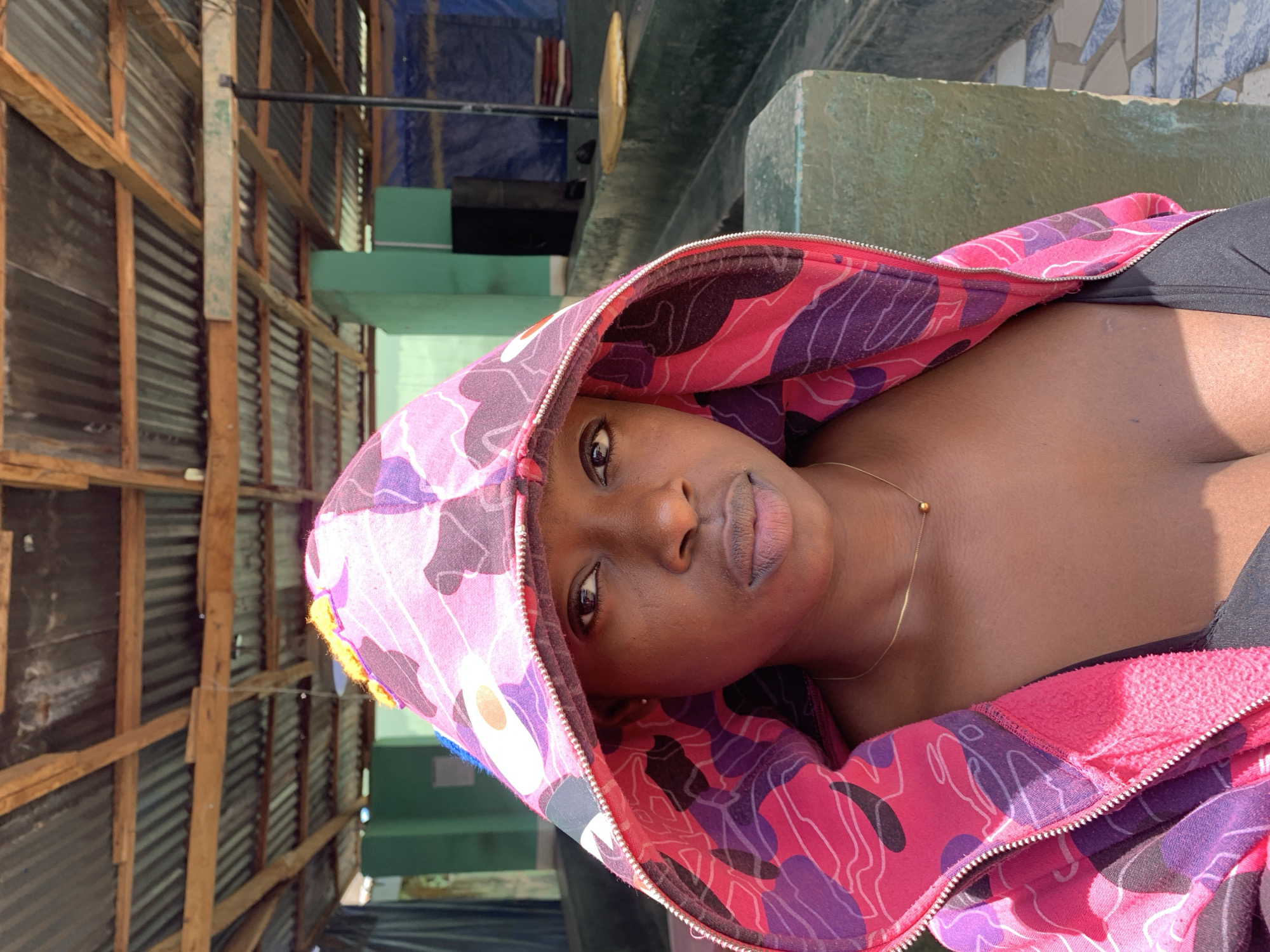 A woman is posing for a photograph in an urban setting, possibly near New Stadium, Nigeria. The woman has dark skin, and is wearing a pink and purple hooded sweatshirt. She is also wearing a thin gold chain around her neck. The background includes a building constructed of wooden planks, and concrete.