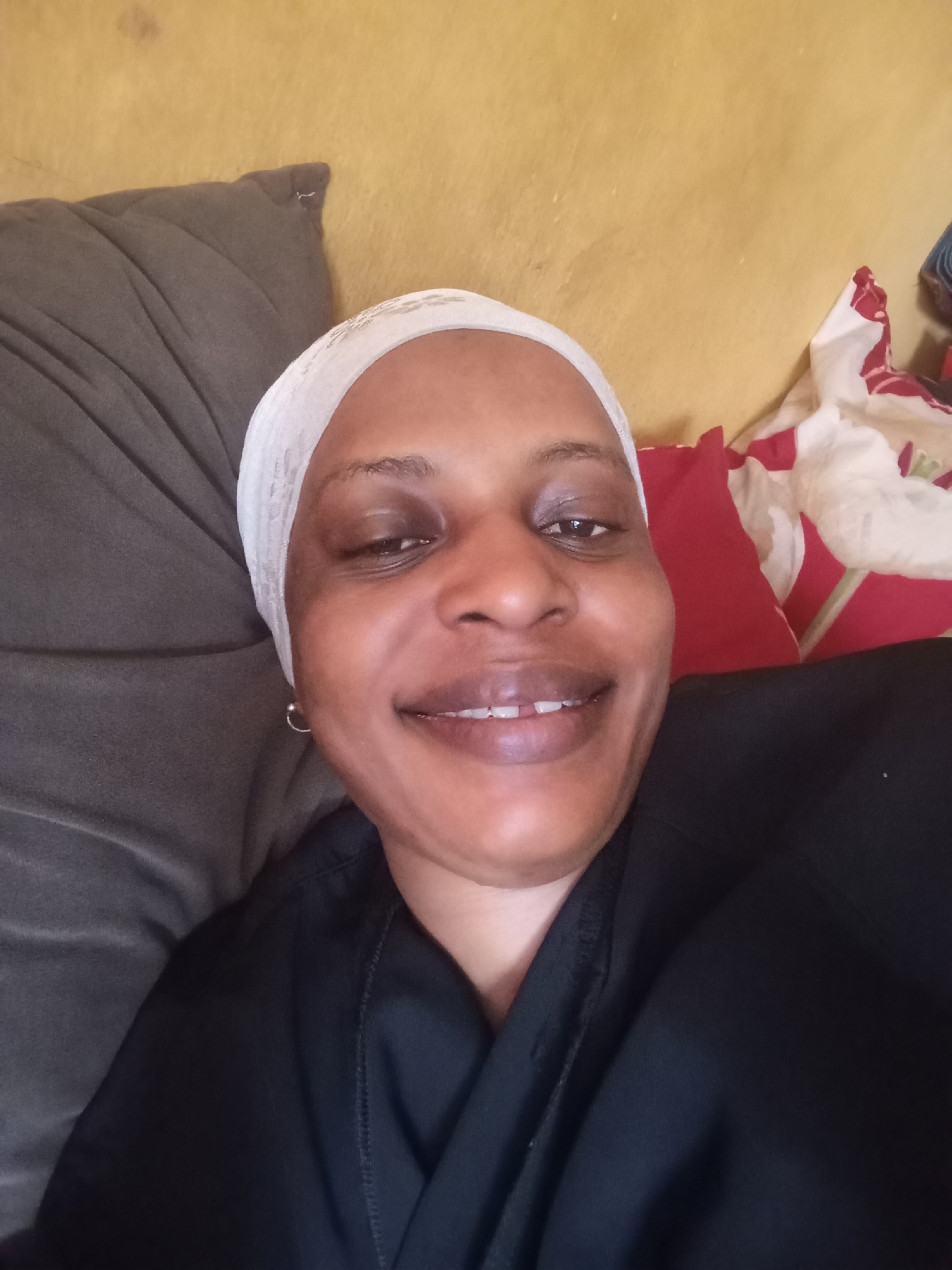 A close-up selfie captures a dark-skinned woman smiling broadly while lying down indoors. She is positioned centrally, her face illuminated by soft, natural light, suggesting it is daytime. Her expression is joyful and friendly, with her eyes open and looking directly at the camera, and a wide smile revealing her teeth, including a slight gap between her front teeth.
The woman wears a white, possibly textured or subtly patterned head covering that frames her face, and a simple black garment, likely a top or dress, covers her body. Small, silver hoop earrings are visible on both ears.
She appears to be resting on a dark grey pillow or cushion, visible on the left side of the frame. To her right, a vibrant red pillow and a white fabric adorned with red patterns, possibly a bedsheet or another cushion, are present. The background consists of a plain, somewhat faded yellow wall, showing minor texture and imperfections, indicating an indoor residential setting. The overall scene conveys a sense of happiness and relaxation. There is no visible text in the image. The setting is a residential indoor space in Narayi, Nigeria.