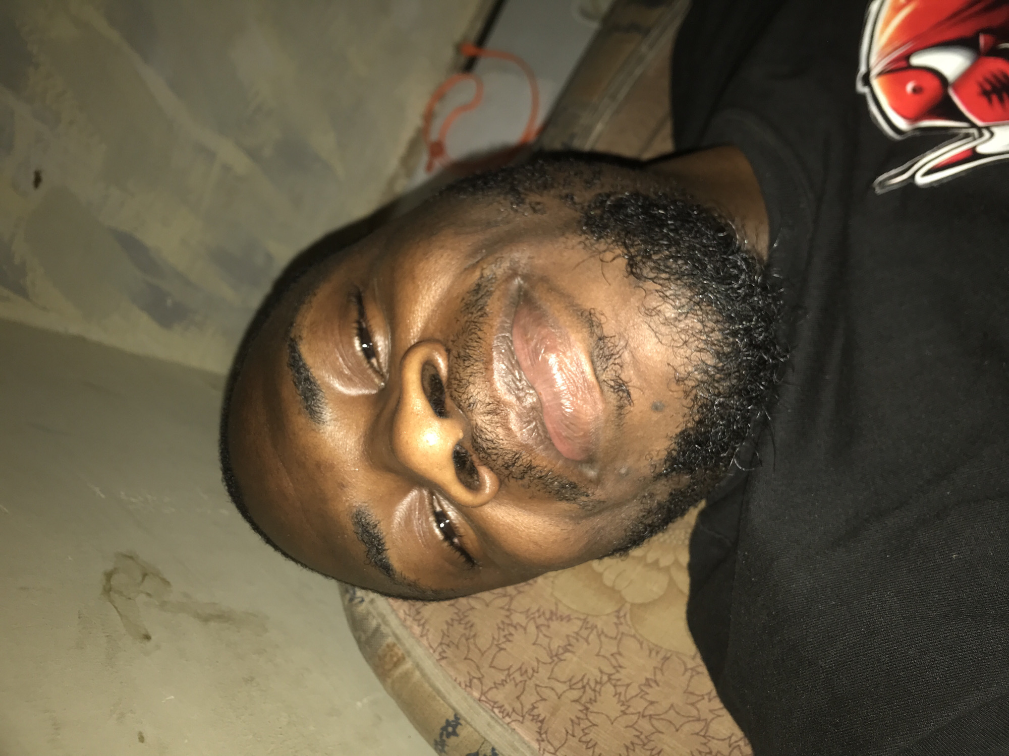 A dark-skinned adult male is depicted in a close-up, head-and-shoulders shot, lying in a supine position and looking directly into the camera. He has short, dark hair, a full, dark, curly beard and mustache. His eyes are open with visible reflections, and his lips are full, slightly parted, and appear moist. He displays a subtle, relaxed facial expression. He is wearing a black short-sleeved t-shirt, on the lower right of which a graphic design with red, white, and black elements, resembling a comic book character's emblem, is partially visible. The subject is resting on a light brown or tan patterned cushion or pillow with darker floral or leaf-like designs. Behind his head, a wall or ceiling is visible, displaying a light beige or off-white base color with areas of apparent discoloration, peeling paint, or water stains, particularly a distinct irregular shape on the upper left. Another section of the background shows vertical light and dark streaks or stripes. A thin, orange, flexible object, possibly a cord or band, is loosely coiled or shaped into an irregular loop on the right side of the background. The scene is illuminated by an indirect light source, creating highlights on the subject's face. The overall environment appears to be an indoor setting in the city of Unknown, Unknown.