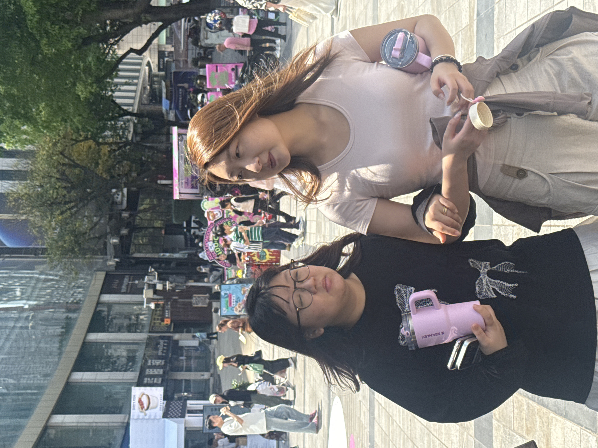 The image depicts two women in the foreground, standing outdoors in a lively urban commercial district during daytime. The scene is bright and clear, suggesting good weather. The location appears to be a bustling pedestrian area with shops and market stalls.
The woman on the right (when oriented correctly) has long, reddish-brown hair and is wearing a light-colored, possibly pale pink, short-sleeved top. She holds a pink insulated tumbler and a small white cup in her hands, and wears a dark beaded bracelet on her left wrist. She looks towards the left with a gentle, slightly smiling expression.
Standing next to her, closer to the viewer, is another woman with dark hair and glasses. She is dressed in a black long-sleeved top adorned with a distinctive white, possibly embroidered, bow-like or winged pattern. She holds a light pink insulated tumbler, clearly marked with the "STANLEY" brand name, in her right hand, and two smartphones in her left hand. Her eyes appear partially closed, perhaps squinting due to the bright light, and she maintains a neutral expression.
The background reveals a vibrant street scene populated by many other pedestrians. Buildings with modern glass facades and commercial signage line the thoroughfare. Green trees are interspersed among the structures. Several market-like stalls or booths, some featuring bright pink decorations, are visible, indicating a commercial or event-filled environment. Distant signs display partially legible text such as "FESTIVAL E", "DOOT", and "DIRTY FISH" accompanied by an image. The overall impression is that of a casual outing in a bustling city setting, likely in Yuzhong District, China.