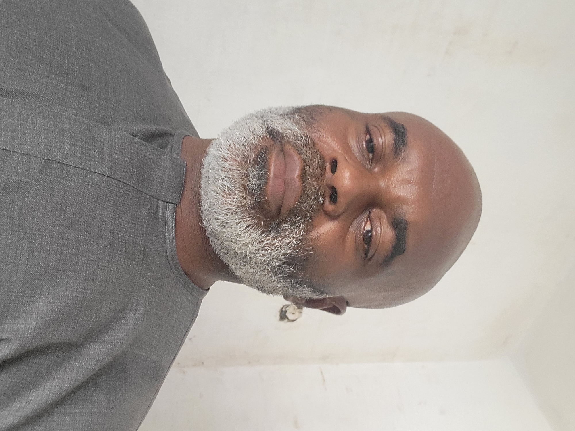 A man with dark skin, a bald head, and a full, grey and white beard is captured in a portrait against a plain, light-colored wall. He is wearing a grey garment, possibly a traditional tunic, with a simple placket neckline. The man looks directly at the camera with a calm, neutral expression. His eyes are dark brown. The background wall appears to be off-white or light beige and shows some slight discoloration and texture. A small, indistinct object or mark is visible on the wall behind his right ear. The lighting suggests an indoor setting, but the time of day and weather are not discernible. No text is visible in the image. The man's appearance and attire are consistent with the provided location context of Maiburiji, Nigeria.