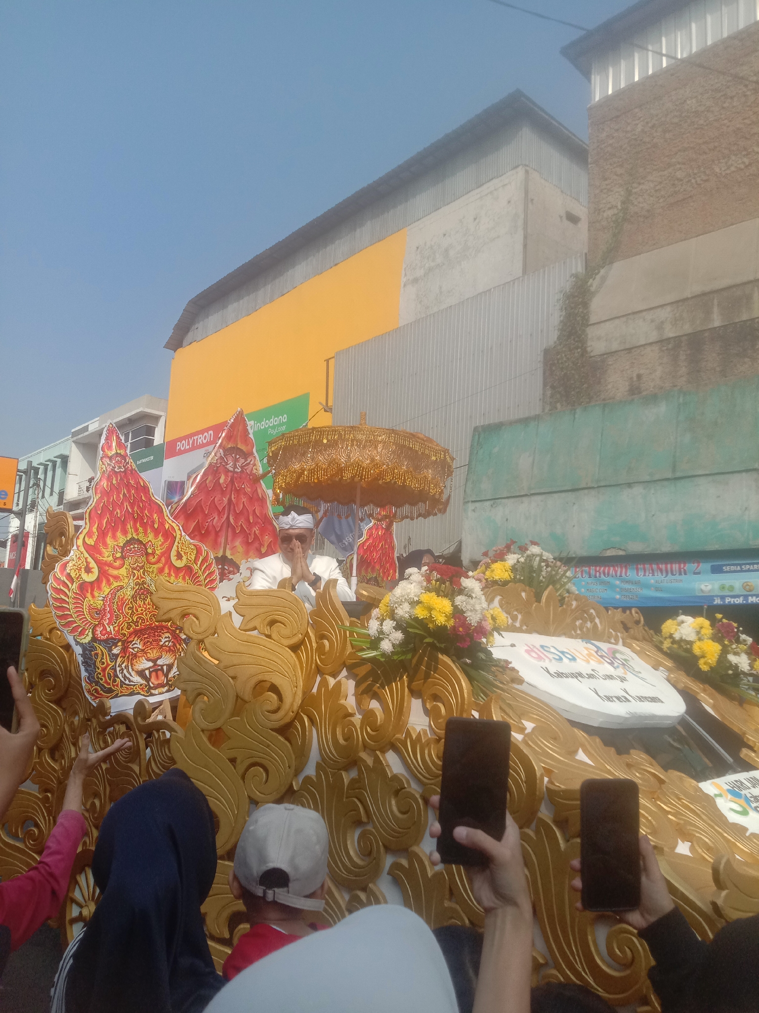The image captures a festive procession in Cianjur, Indonesia. In the foreground, several people are holding up smartphones, presumably to capture the event. The main focus is a decorated float adorned with golden, ornate carvings and vibrant floral arrangements. A man in traditional attire, wearing a white head covering, is seated on the float, with his hands clasped in a gesture of greeting or prayer. Behind him, there are large, stylized cutouts featuring a tiger and fiery motifs. In the background, buildings are visible, including one with a bright yellow facade and another with a textured, brick-like exterior. A sign that reads "ELECTRONIC CJANJUR 2" is also present, hinting at the procession's location within the city. The overall atmosphere appears to be that of a public celebration or parade.