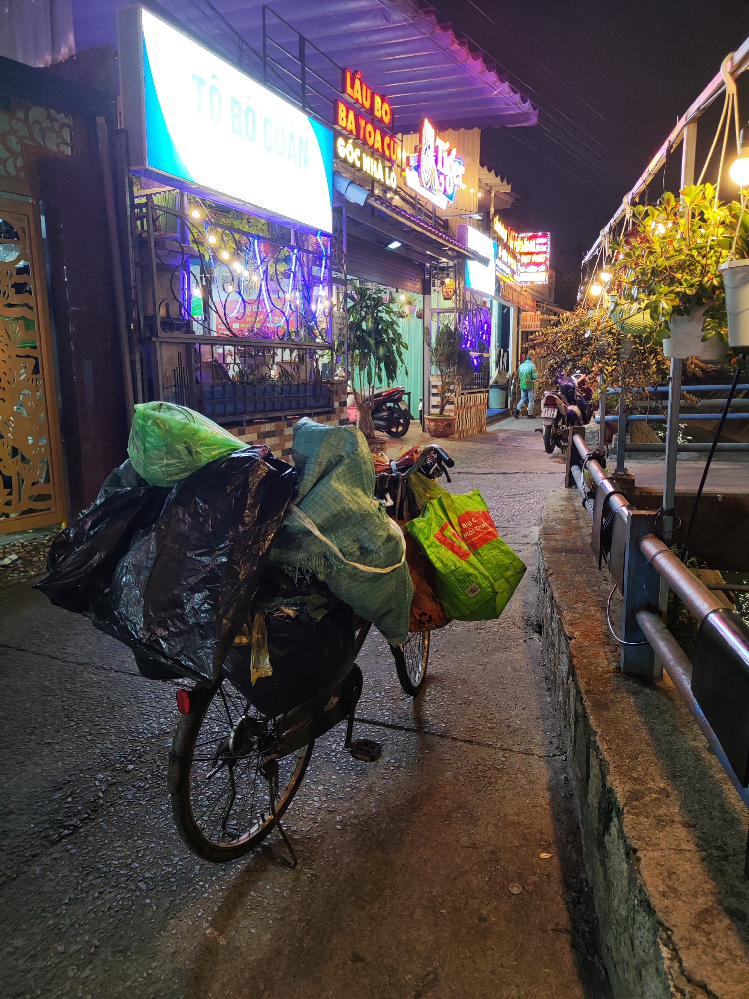 A bicycle overflowing with bags, likely used for transporting items, stands on a street in Cam Ly - Đà Lạt, Vietnam, at night. The bicycle is positioned on a narrow sidewalk next to a low concrete wall with a metal railing. Behind the bicycle, the street is lined with businesses, identifiable by their illuminated signs, including "TO BO QUAN", "LAU BO BA TOA CU", and "HU TIEU AU DA LAT". The bags on the bicycle are a mix of black plastic and woven materials, with a green shopping bag from "Big C" visible on the side. A motorcycle is parked further down the street, and a person can be seen walking away in the distance. The street is lit by the storefronts and hanging light fixtures with plants.