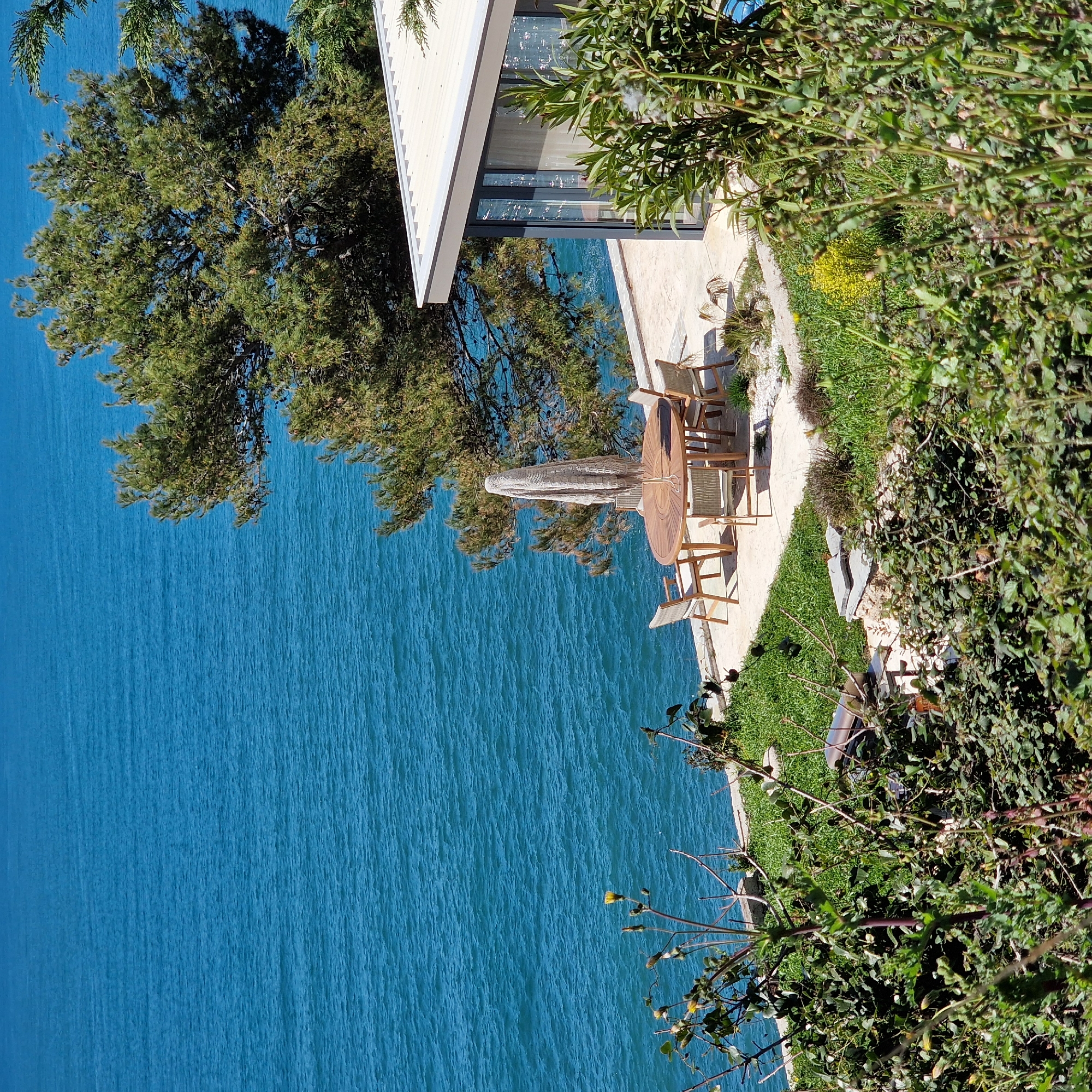 A high-angle view captures a waterfront scene. To the left, vibrant turquoise water dominates, merging with the sky on the horizon. Adjacent to the water is a property featuring a modern architectural style. A patio area is furnished with a round wooden table and chairs, sheltered by a white umbrella. Dense greenery, including trees and shrubs, frames the right side of the view. The scene is located in the city of Ulcinj - Ulqin, Montenegro.