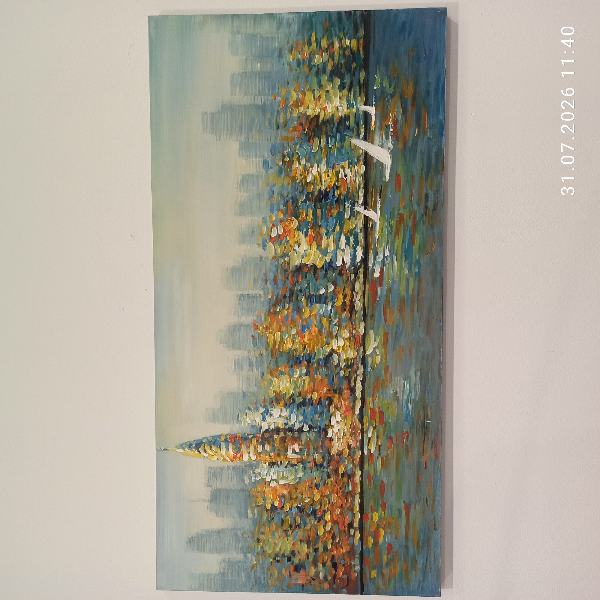 This is a rectangular painting of a city skyline at dusk. The city is depicted with tall buildings in shades of gray and blue, with a prominent, colorful skyscraper in the center that is painted with thick, impasto strokes of yellow, orange, red, blue, and white. The water in the foreground reflects the city lights in a similar palette of vibrant colors. Three sailboats with white sails are visible on the water, moving from left to right. The painting is hanging on a textured, off-white wall. A timestamp "31.07.2026 11:40" is overlaid on the bottom right corner of the image. The location of Modugno, Italy is not depicted in the painting.
