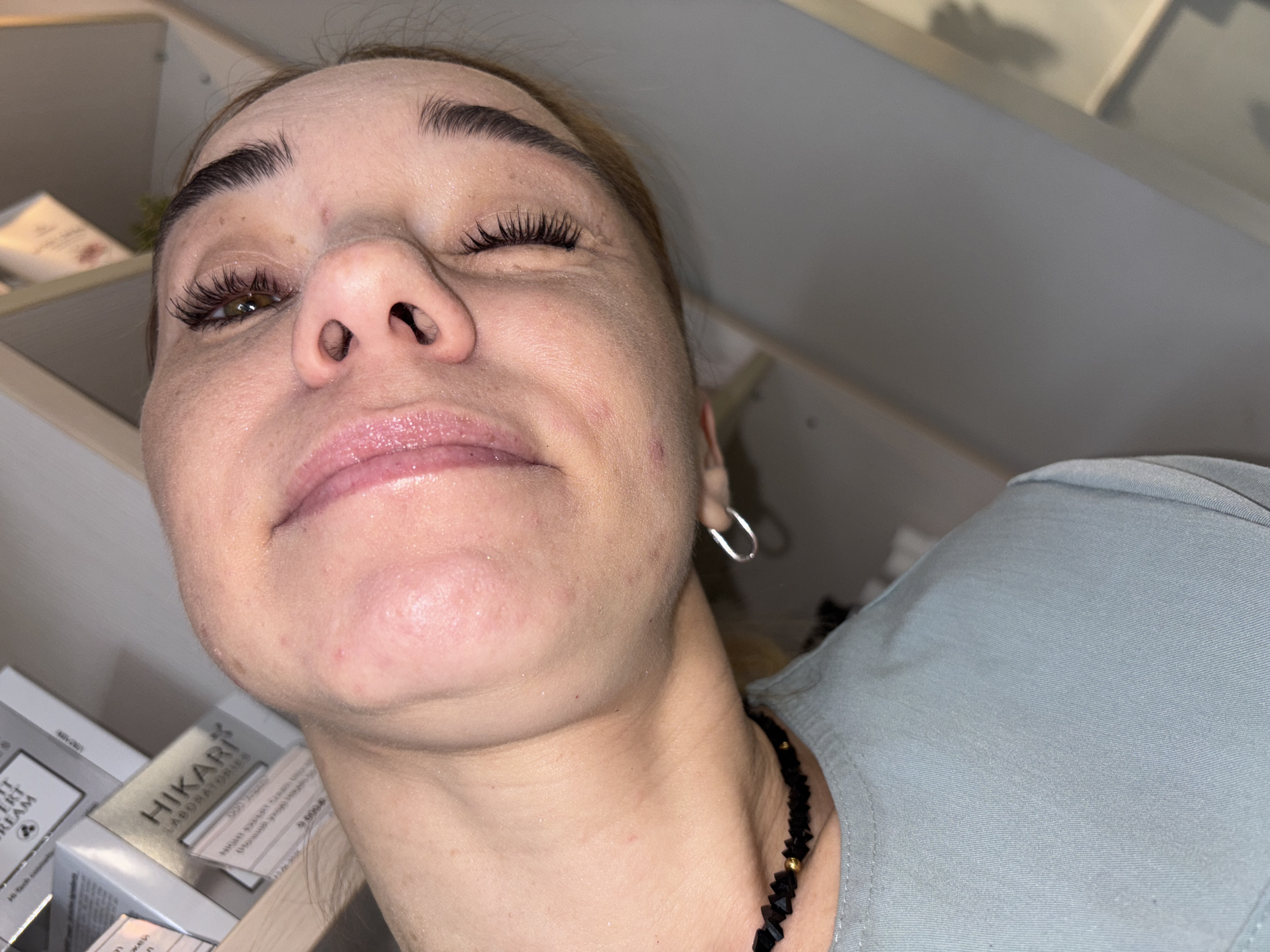 The image shows a close-up, slightly tilted, overhead view of a person's face and upper chest. The person appears to be female, with fair skin, dark eyebrows, and visible eyelashes. Her mouth is slightly open, revealing her tongue. She is wearing a light blue top and a black beaded necklace with gold accents. A silver hoop earring is visible on her right earlobe. In the background, shelves are visible with boxes of cosmetic products. The environment appears to be indoors, possibly a beauty salon or retail space. The specific location in Ladozhskoe Ozero, Russia cannot be determined from the image alone, as there are no identifying landmarks or context clues to suggest this location.
