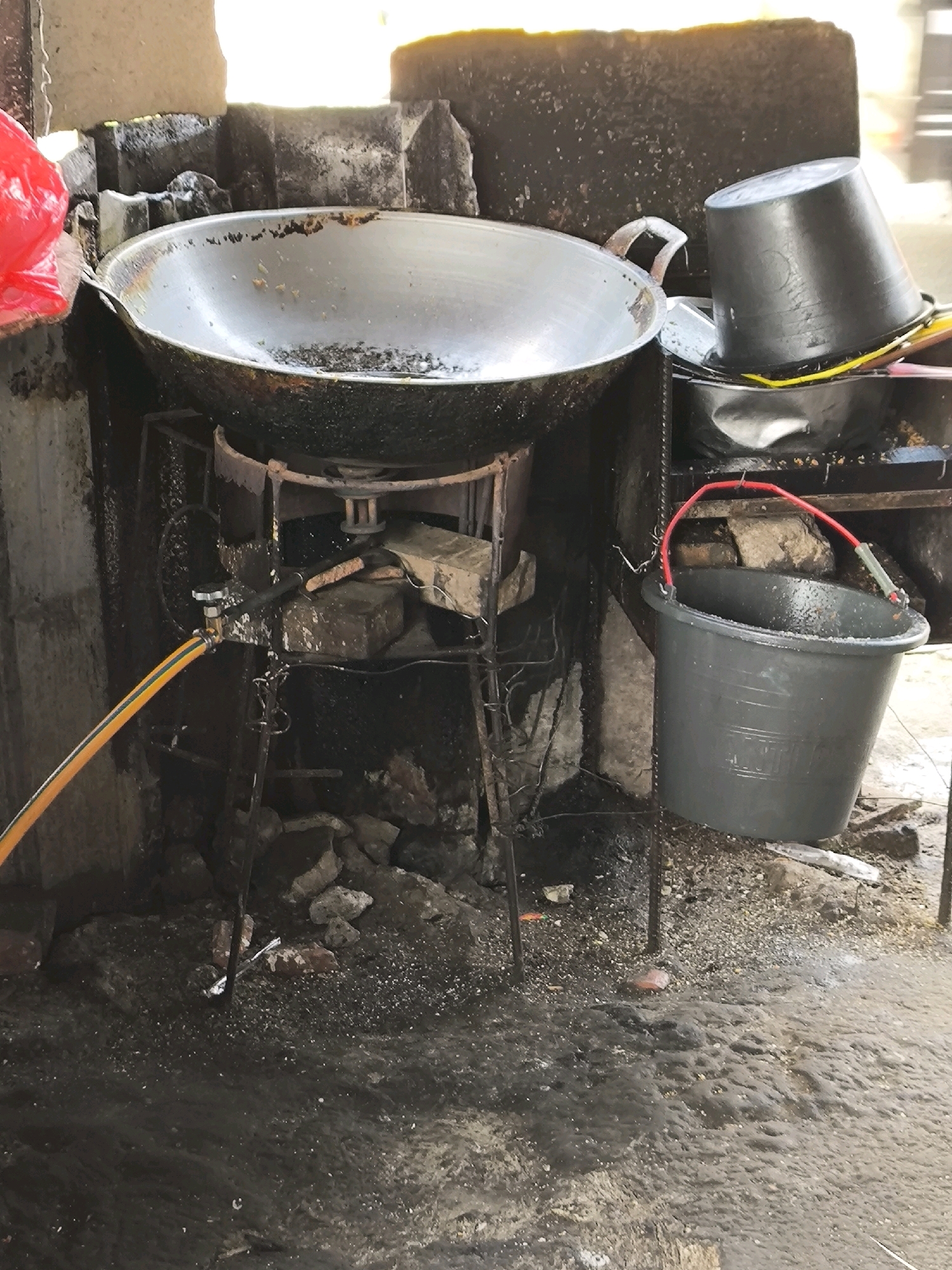 The image depicts an outdoor scene at a food stall in Lubangbuaya, Indonesia. A large, well-used wok rests on a metal stand above what appears to be a makeshift stove constructed from bricks. To the right of the wok, a black plastic bucket is stacked on top of another darker container, with a yellow hose visible between them. Below the stove, a gray plastic bucket with a red handle hangs from a wire. An orange and yellow striped hose is attached to a metal fixture on the left side of the stove, extending out of frame. The ground is wet and uneven, covered with debris including rocks and what appear to be small pieces of white material. The overall environment suggests a functional but unkempt preparation area. There are no people visible in the frame.