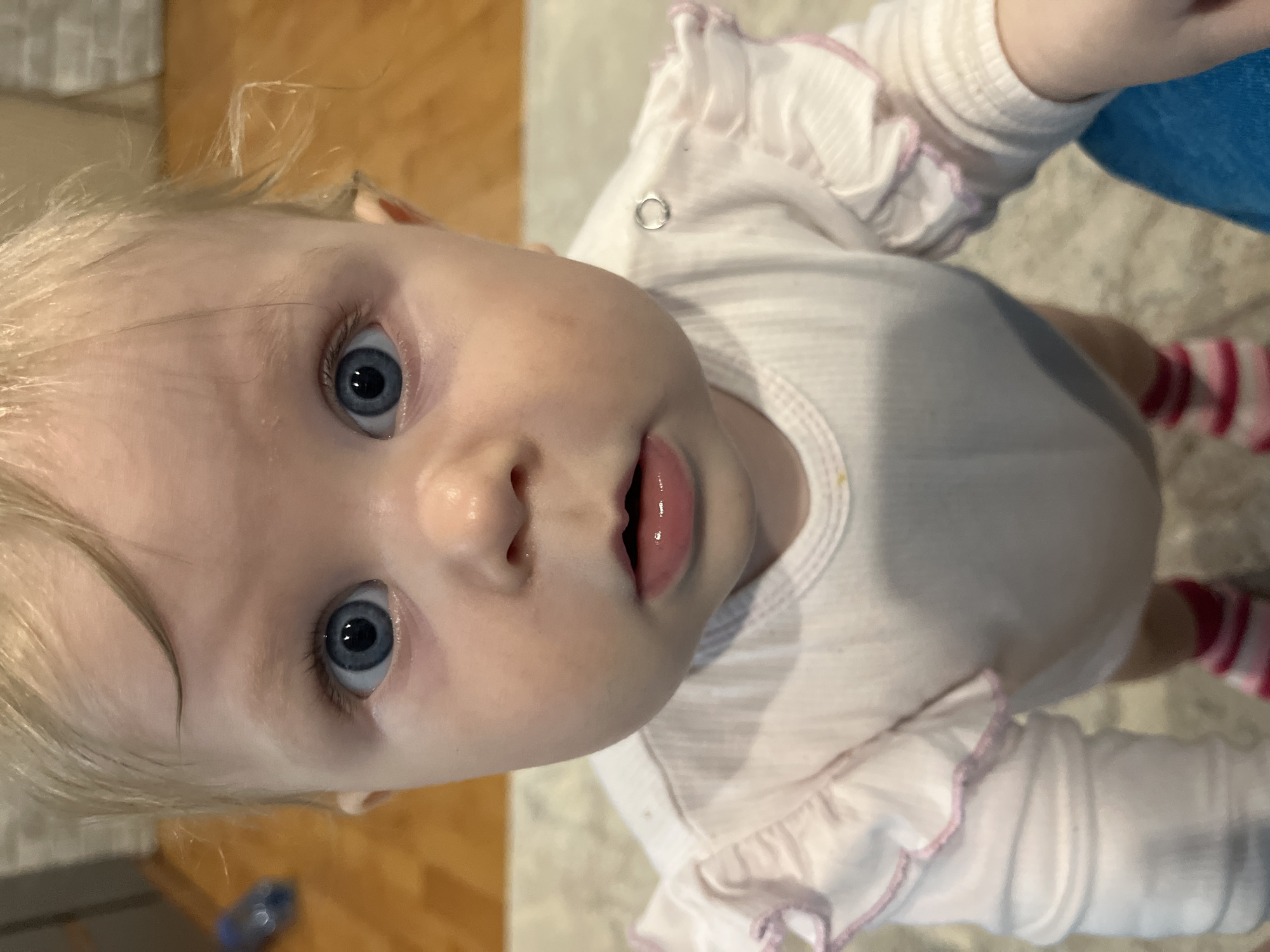 A high-angle, close-up photograph shows a baby with blonde hair and blue eyes looking directly at the camera. The baby is wearing a white onesie with pink trim on the sleeves. The lower legs are visible with striped pink and white socks. Part of a blue garment is visible on the right side. The background includes a cream-colored surface with a wood-like pattern, and another surface with a different wood-like pattern in the lower left corner, along with a blue object on the ground. The location is not specified.
