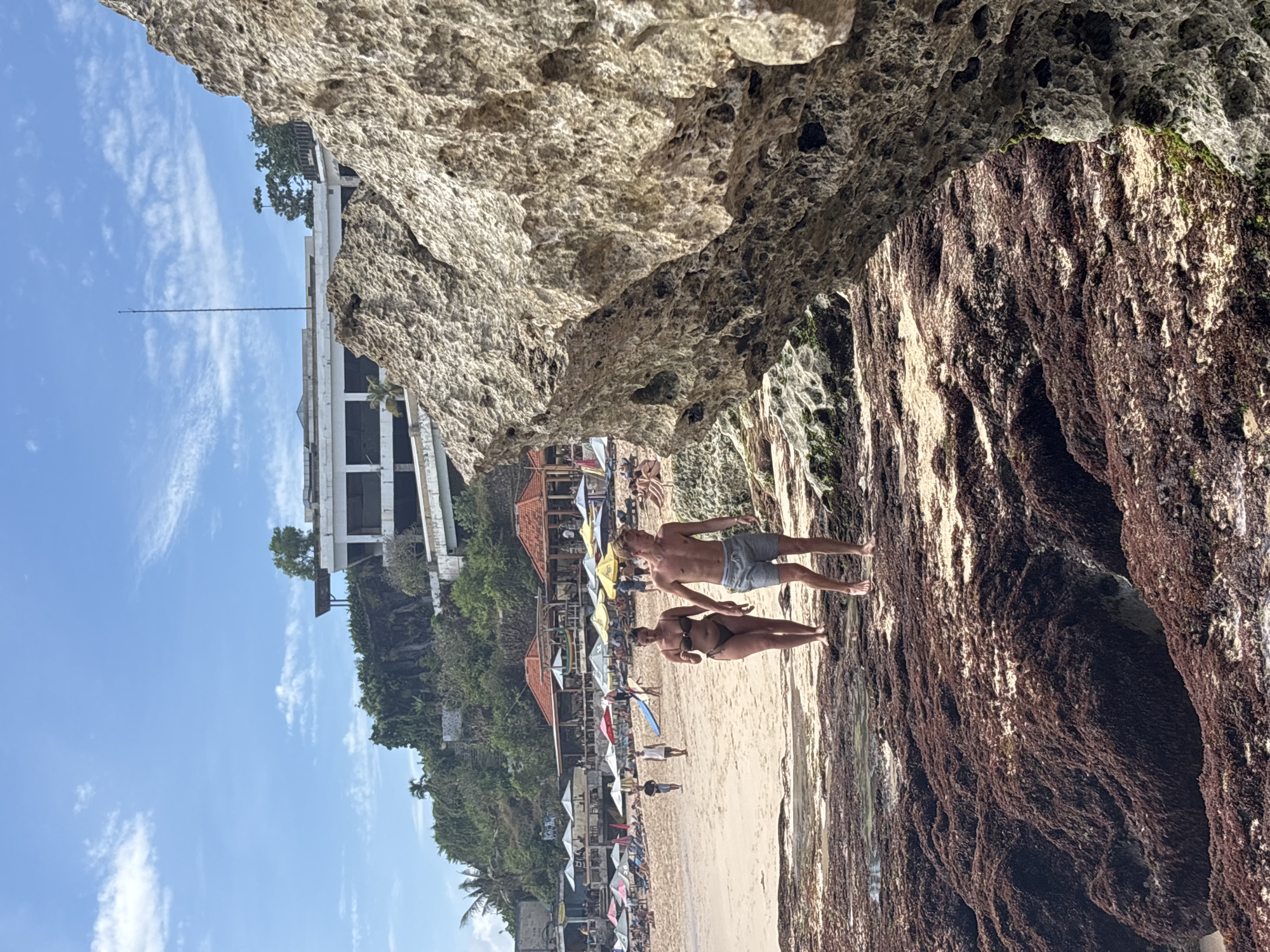 In Kuta Selatan, Indonesia, this image captures a beach scene under a bright blue sky with scattered clouds. In the foreground, a large, textured rock formation dominates the right side of the frame, partially obscuring the view of the beach. Two individuals, a woman in a dark bikini and a man in grey swim trunks, walk barefoot on the wet, rocky shore in the mid-ground. The beach stretches out behind them, lined with numerous colorful umbrellas and lounge chairs, suggesting a popular tourist destination. Several beachgoers are visible, some relaxing on the sand and others walking along the shoreline. In the distance, a multi-story building with a white facade is perched on a green, vegetated cliff overlooking the beach. Further up the cliff, more buildings and foliage are visible, with a tall antenna rising into the sky. The overall scene depicts a vibrant and active beach resort area on a sunny day.