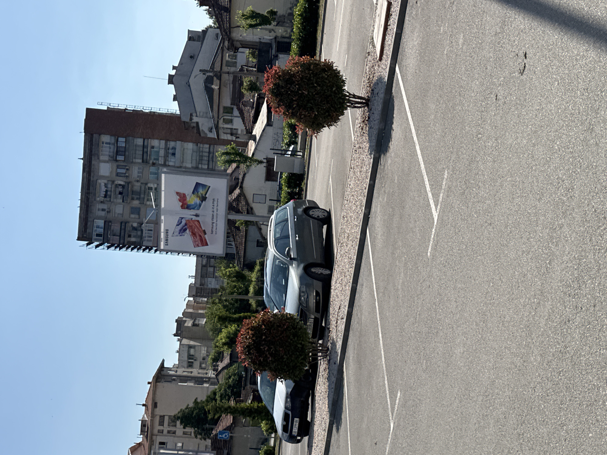The image depicts a parking lot in Lovech, Bulgaria, lined with two cars parked in designated spaces. The vehicles appear to be a dark-colored sedan, followed by a silver car. Small, rounded bushes separate the parking spots. A building with a large Samsung advertisement on its side stands in the background, along with other residential buildings. The sky is clear and blue. The scene is well-lit, suggesting it is daytime.