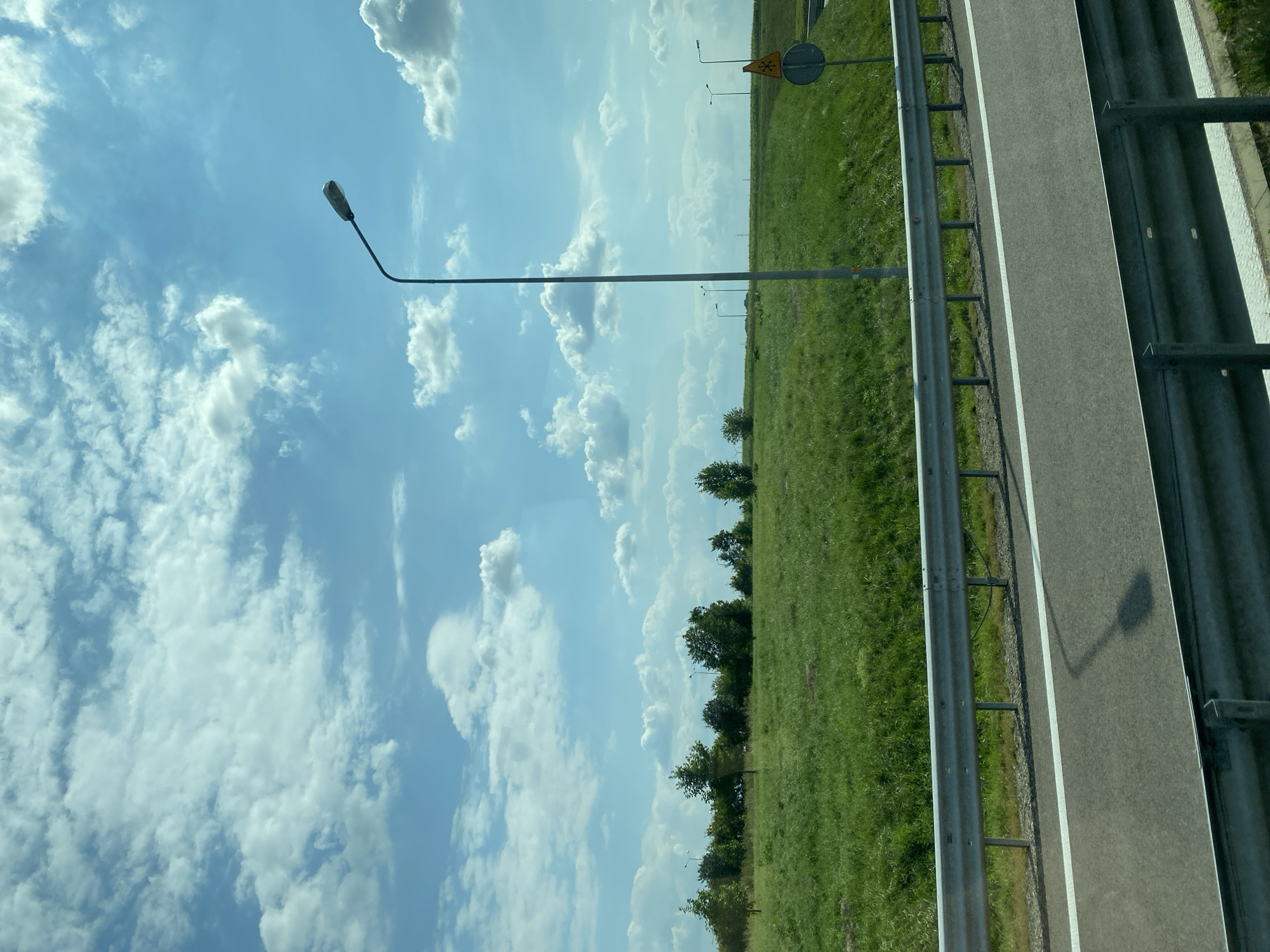 The image shows a bright, partly cloudy sky over a grassy field. A tall street lamp stands on the right side of the frame, casting a shadow on the asphalt road in the foreground. A metal guardrail separates the road from the field, which is dotted with trees. A yellow traffic sign with a snowflake symbol is visible near the street lamp. The scene appears to be a roadside in or near Skołoszów, Poland.
