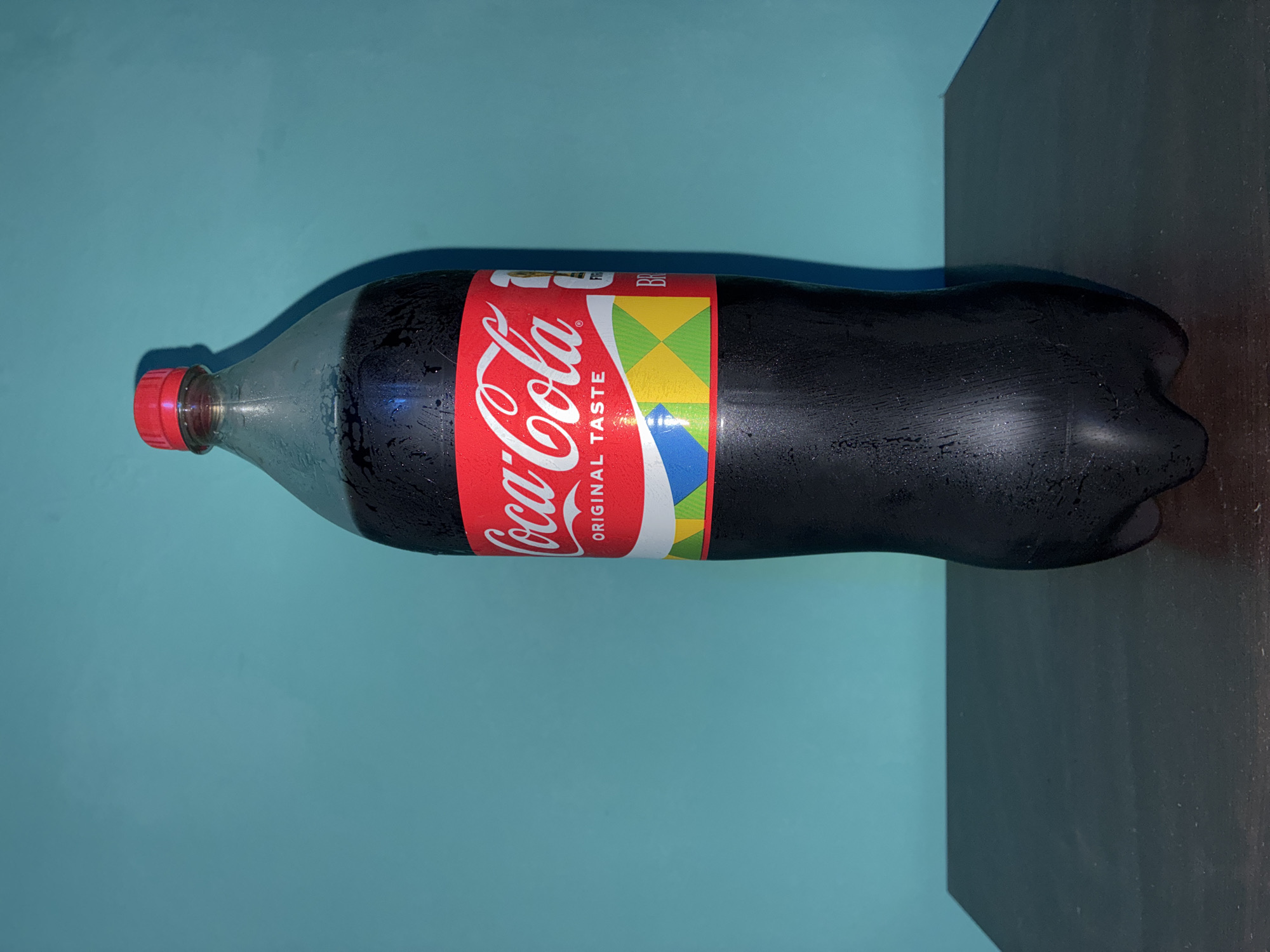 A horizontal, medium shot shows a 2-liter plastic bottle of Coca-Cola. The bottle is mostly full and has condensation on its surface, indicating it is likely cold. The cap is red, and the Coca-Cola label features the iconic red and white logo with the words "Original Taste." A portion of the label has geometric shapes in colors of green, yellow, and blue. The bottle rests on a dark gray or black surface, possibly a table or shelf. The background is a solid turquoise color, suggesting a wall. The scene appears to be indoors. I am unable to determine from the image if it is located in Elhovo, Bulgaria.