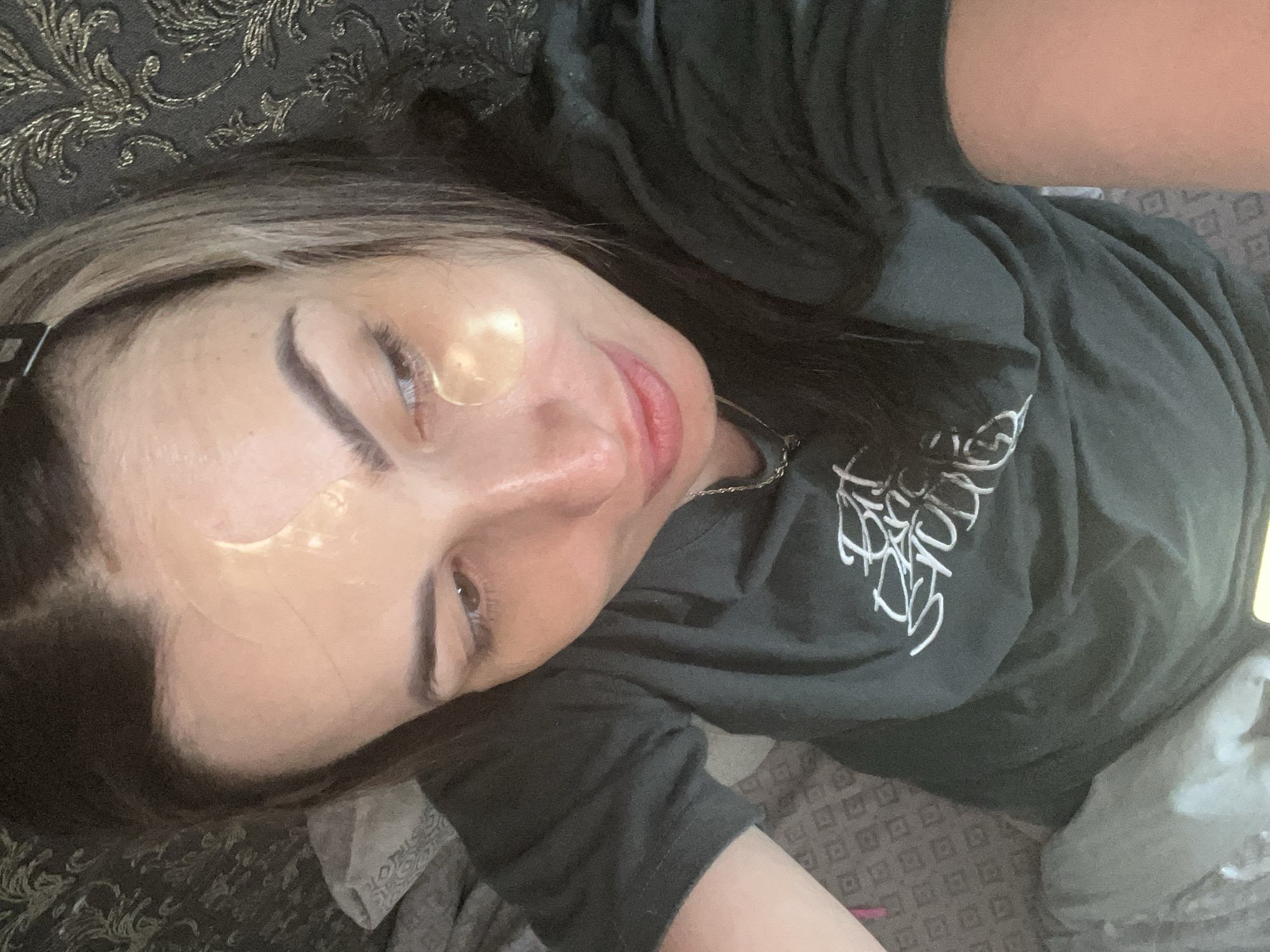 A close-up selfie captured from an overhead angle shows a person, likely female, lying down indoors. The individual is positioned diagonally across the frame, looking towards the camera with a gentle, relaxed expression and a soft smile. They have dark hair, some strands of which appear lighter, possibly with highlights, pulled back with a dark hair clip visible on the left side of their head. Two clear, circular or crescent-shaped hydrogel under-eye patches are visible on their face, one below each eye.
The person is wearing a dark, possibly charcoal or black, short-sleeved t-shirt with white, stylized text printed on the front. The text is partially visible but appears to read "Eat Sleep Build Repeat". A silver-colored chain necklace is also visible around their neck.
The background consists of patterned fabrics, suggesting an indoor setting like a bedroom or living room. Behind the person's head, a dark fabric with intricate gold embroidered or woven patterns is visible, likely a pillow or headboard. Below their arm and to the right, another fabric with a repetitive square or diamond geometric pattern in muted tones can be seen. The overall impression is one of a casual, relaxed moment, possibly during a skincare routine or unwinding.