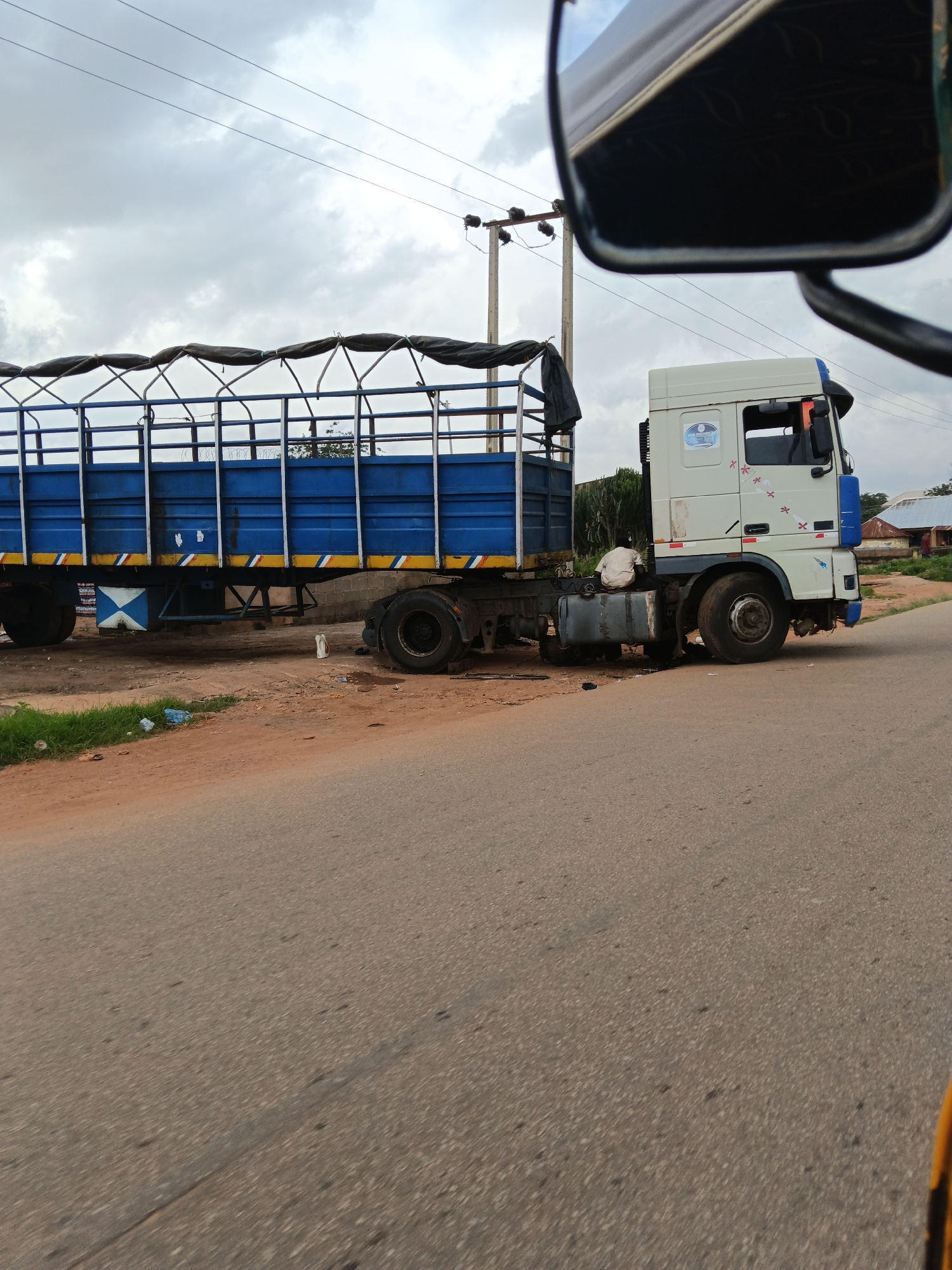 A semi-truck with a blue trailer is parked on the side of a road. The truck is white with blue accents and has a logo on the side of the cab. The trailer is open-topped and covered with a dark tarp. A person in a white shirt is sitting on a fuel tank on the side of the truck, facing away from the camera. There are power lines and poles in the background, along with some buildings and vegetation. The road is paved, and there is a dirt shoulder with some debris. The sky is overcast.