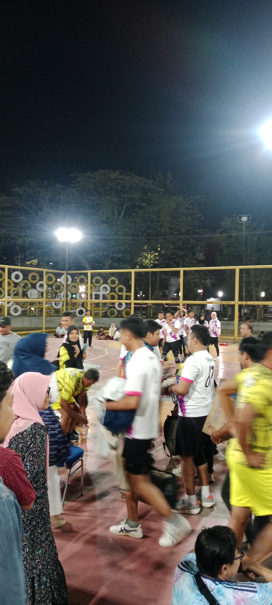 A crowd of people are gathered on a pink sports court at night in the city of Tuban, Indonesia, likely at GOR (Gedung Olah Raga) Anung. The scene is illuminated by bright overhead lights. Many individuals are wearing athletic attire, with some in matching white shirts featuring pink accents and numbering, suggesting a team or event. One person in the foreground wears a white shirt with the number "87" visible. Other people are dressed in casual clothing, including a woman in a pink hijab and a person in a blue hijab. Some individuals appear to be carrying bags or equipment. A yellow fence with circular patterns, possibly made of tires, encloses part of the court. Trees are visible in the background, silhouetted against the night sky. The overall atmosphere suggests an informal sporting gathering or event.