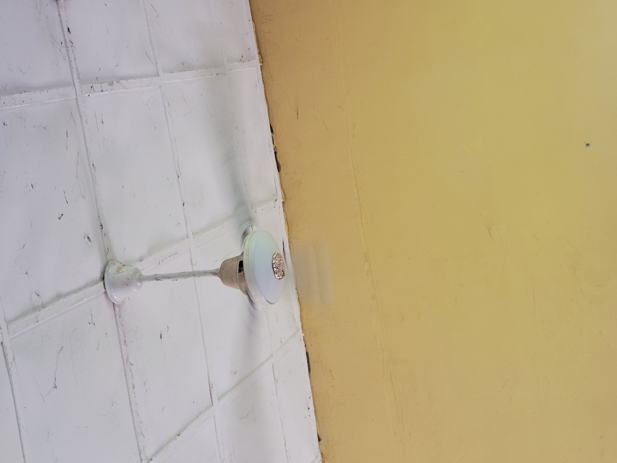 An indoor shot shows a ceiling fan suspended from a white, tiled ceiling. The ceiling appears to have some age and minor imperfections. Below the ceiling is a yellow-painted wall. The image composition focuses on the fan and the upper portion of the wall. The scene is likely within a residential or public building in Keffi, Nigeria, given the visual context of the fan, ceiling, and wall finish.
