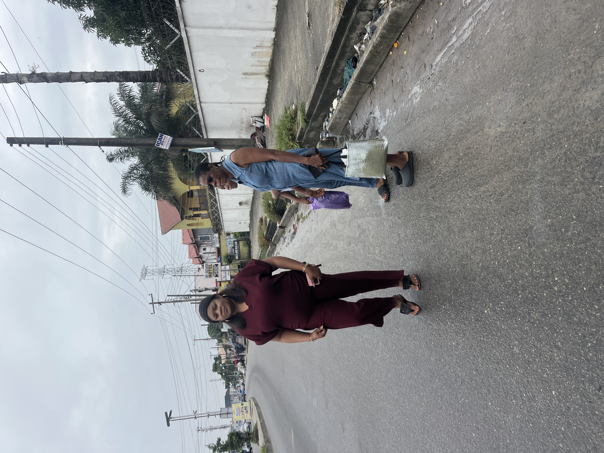 Three women are walking on a wet asphalt road in Ado, Nigeria. The woman on the left is wearing a maroon jumpsuit and black flip-flops, carrying a pink cell phone. The middle woman is wearing a denim jumpsuit and black slides, carrying a purple bag and a black clutch. The woman on the right is also wearing black slides and is carrying a large clear bag. The background shows a street with buildings, power lines, and palm trees. There is a white wall with barbed wire along the sidewalk, and trash is visible along the gutter. The sky is overcast.
