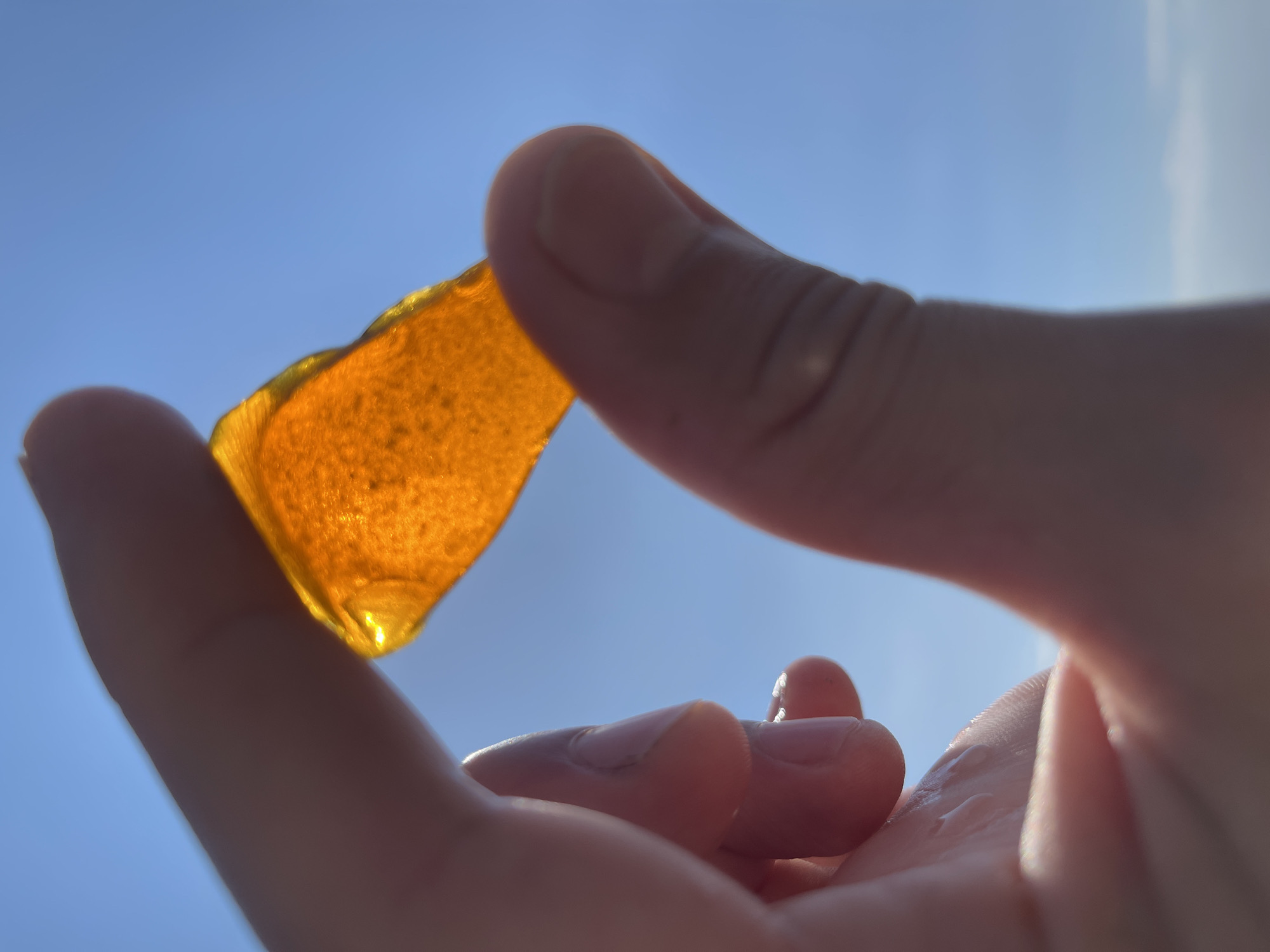 A close-up, low-angle shot captures a person's hand holding a piece of translucent amber-colored sea glass against a clear blue sky. The sea glass is irregular in shape and shows small imperfections. The fingers are slightly wet, with light reflecting off the moisture. The background is dominated by the bright, cloudless sky, indicating an outdoor, likely coastal, setting. The image does not provide any specific details that would allow for the determination of its location within the city of Zatoka, Ukraine.