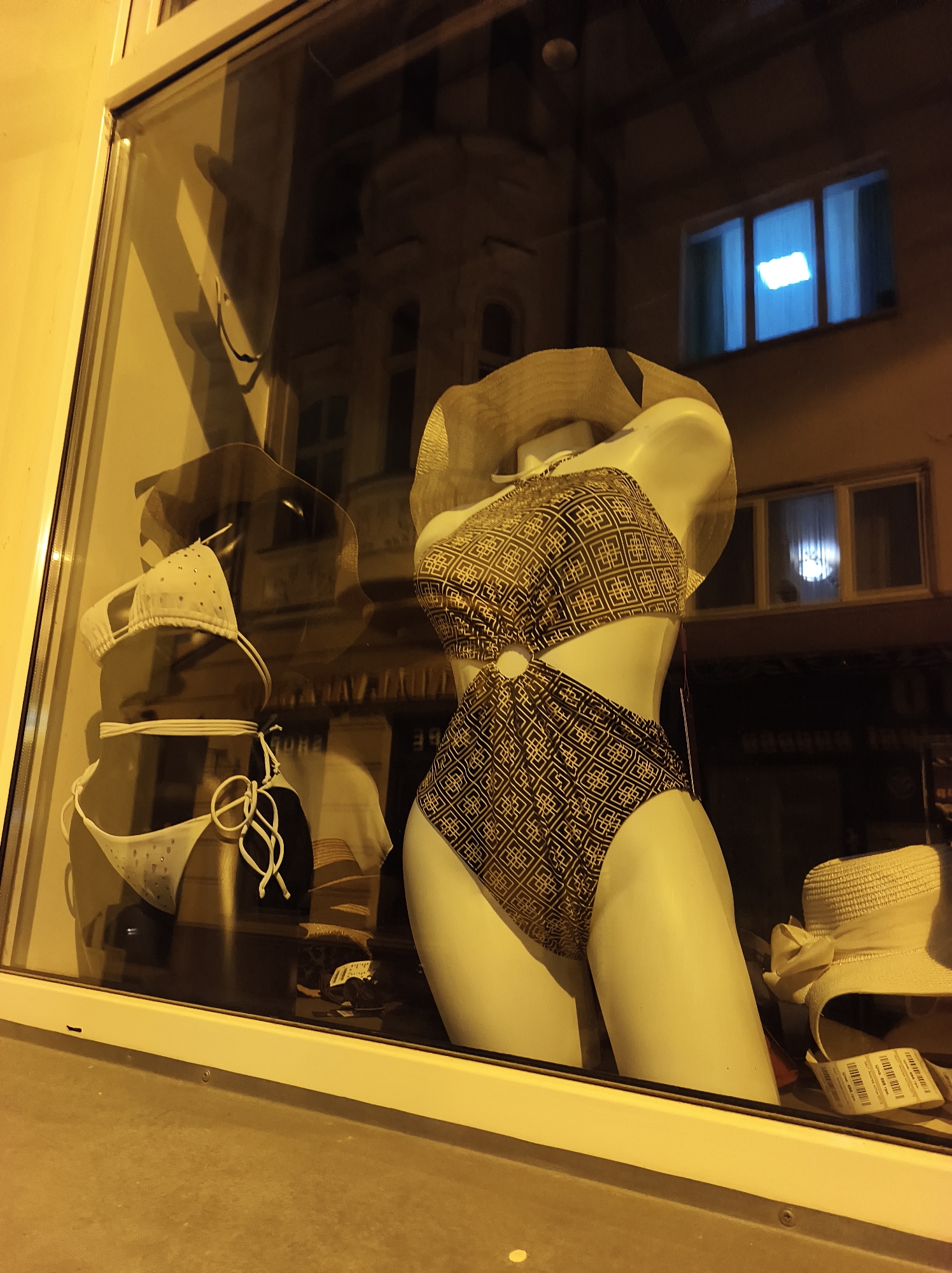 The image shows a storefront window at night in Ternopil, Ukraine. Two mannequins wearing swimsuits are displayed. One mannequin, on the right, is wearing a dark swimsuit with a geometric pattern and a straw hat. Another mannequin, on the left, displays a white swimsuit with white ties. Reflections of buildings across the street are visible in the window glass. A white hat is also visible. Tags are visible on the hat.