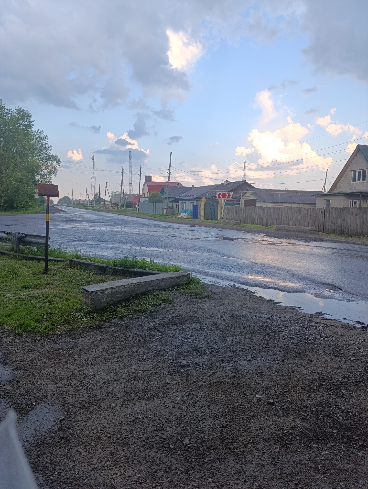 The image depicts a rural road in Tugulym, Russia, after a rainfall. The asphalt is wet and reflects the overcast sky, with several puddles indicating the recent precipitation. The road is lined with grass and gravel on the sides, and a metal guardrail is visible on the left. In the foreground, a concrete structure lies on the grass.
Along the road, houses with wooden fences and various roofing materials are scattered. Some of the houses have visible windows and doorways. In the distance, several telecommunication towers are prominent against the sky. The sky itself is a mix of blue and grey clouds, with hints of sunlight breaking through, suggesting it is either early morning or late afternoon.
A road sign with red and white chevron markings is placed near the intersection. The overall scene conveys a sense of quiet rural life, with no people or animals visible. The weather appears to be transitioning from rain, leaving the environment damp and fresh. The presence of telecommunication towers suggests the area is connected, despite its rural setting. The image is a still photograph, capturing a moment in time in a Russian village.