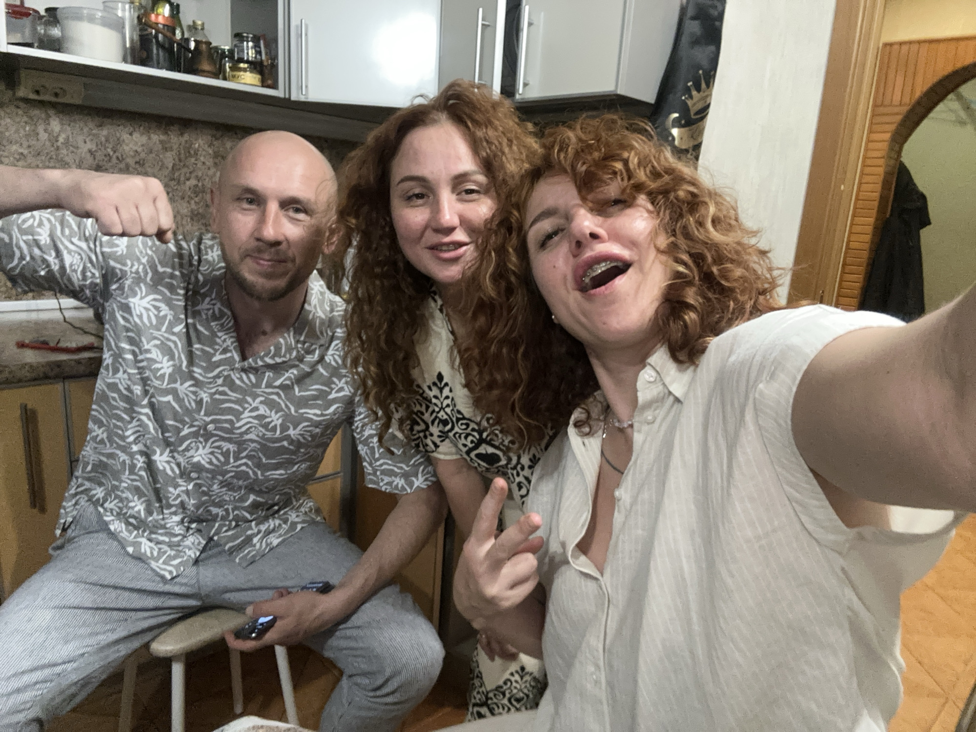 A selfie-style, eye-level photograph taken indoors in a residential kitchen or dining area in Sochi, Russia, depicts three individuals. On the left, a bald man with a short beard, wearing a grey short-sleeved button-up shirt with a white palm frond pattern and grey striped pants, is seated on a light-colored stool. He is facing forward with a slight smile, his right arm bent with a closed fist near his shoulder, and his left hand holding a dark-colored smartphone with an illuminated screen. To his right, a woman with long, curly, reddish-brown hair and light skin, wearing a light-colored top with a black and white geometric pattern, stands slightly behind him, leaning forward with a gentle smile directed at the camera. On the far right, partially cropped, is another woman with curly, reddish-brown hair, light skin, and visible dental braces on her upper teeth. She wears a light-colored, vertically striped button-up shirt and two necklaces, one beaded and one with a dark cord. This woman, positioned as if taking the photograph, has her mouth open in a wide smile and her right hand extended forward, displaying a gesture with her index and middle fingers extended. The background behind the man and the central woman features a textured beige wall, light-colored upper kitchen cabinets with silver handles, and a shelf displaying various containers, bottles, and jars. To the right, a white wall transitions into a dark wooden archway, revealing a partial view of an adjacent room with a mirror and a dark garment hanging.