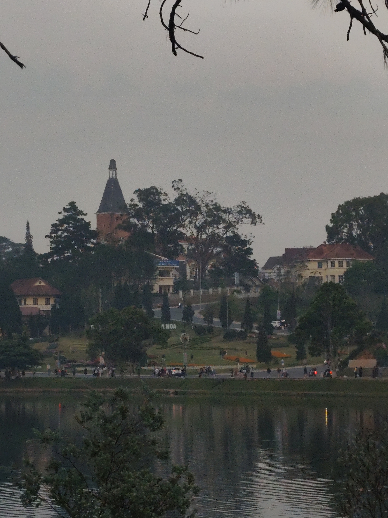 The image depicts a panoramic view of Xuân Hương Lake and its surrounding cityscape in Da Lat, Vietnam, under an overcast sky. In the foreground, the rippled surface of Xuân Hương Lake occupies the lower third, reflecting faint lights from the opposite shore. Dark, out-of-focus tree branches partially frame the top left and top right corners of the image.
Above the lake, a paved road runs horizontally, populated by numerous pedestrians and several motorcycles/scooters, some with riders. Adjacent to the road is a landscaped park area featuring green grass, trees, and a large white sign partially displaying "T. HOA". A white car is visible on a secondary road further back in the park.
On the hillside behind the park and road, several buildings are interspersed among dense trees. The prominent structure is the Da Lat Railway Station, identifiable by its reddish-brown base, tall, dark conical roof, and lighter cylindrical top, positioned towards the center-left. Other buildings include a multi-story structure with a reddish-brown tiled roof and light-colored facade, and a white building with blue roof accents. The sky above is uniformly light gray and overcast.