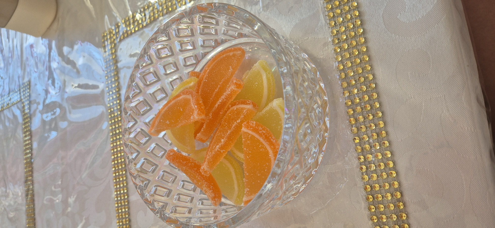 A crystal bowl filled with orange and lemon slice candies sits on a white patterned tablecloth with gold trim.